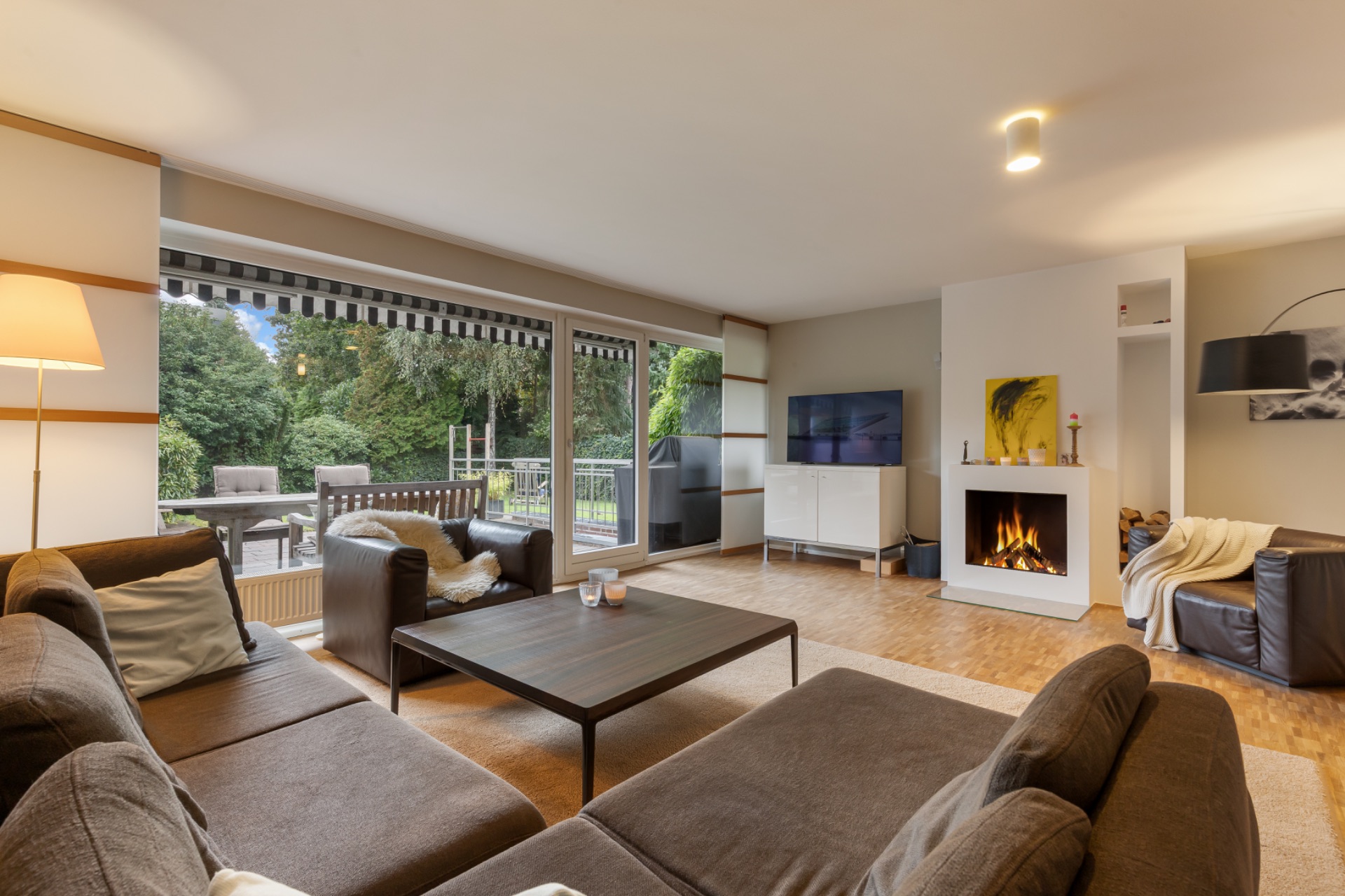 Modernes Wohnzimmer mit Kamin, Sofa, TV und großer Fensterfront zum Garten.