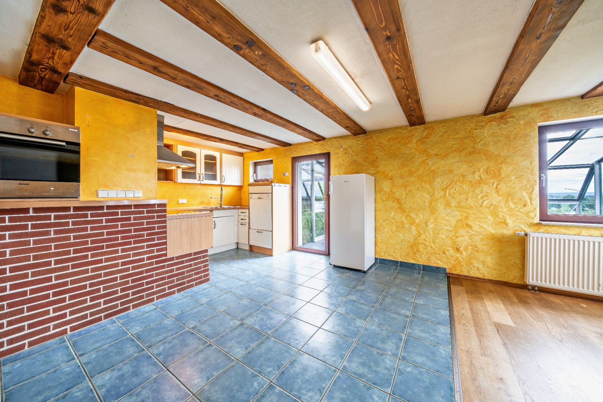 Helle Küche mit gelben Wänden, Holzbalken, blauen Fliesen und großem Fenster.