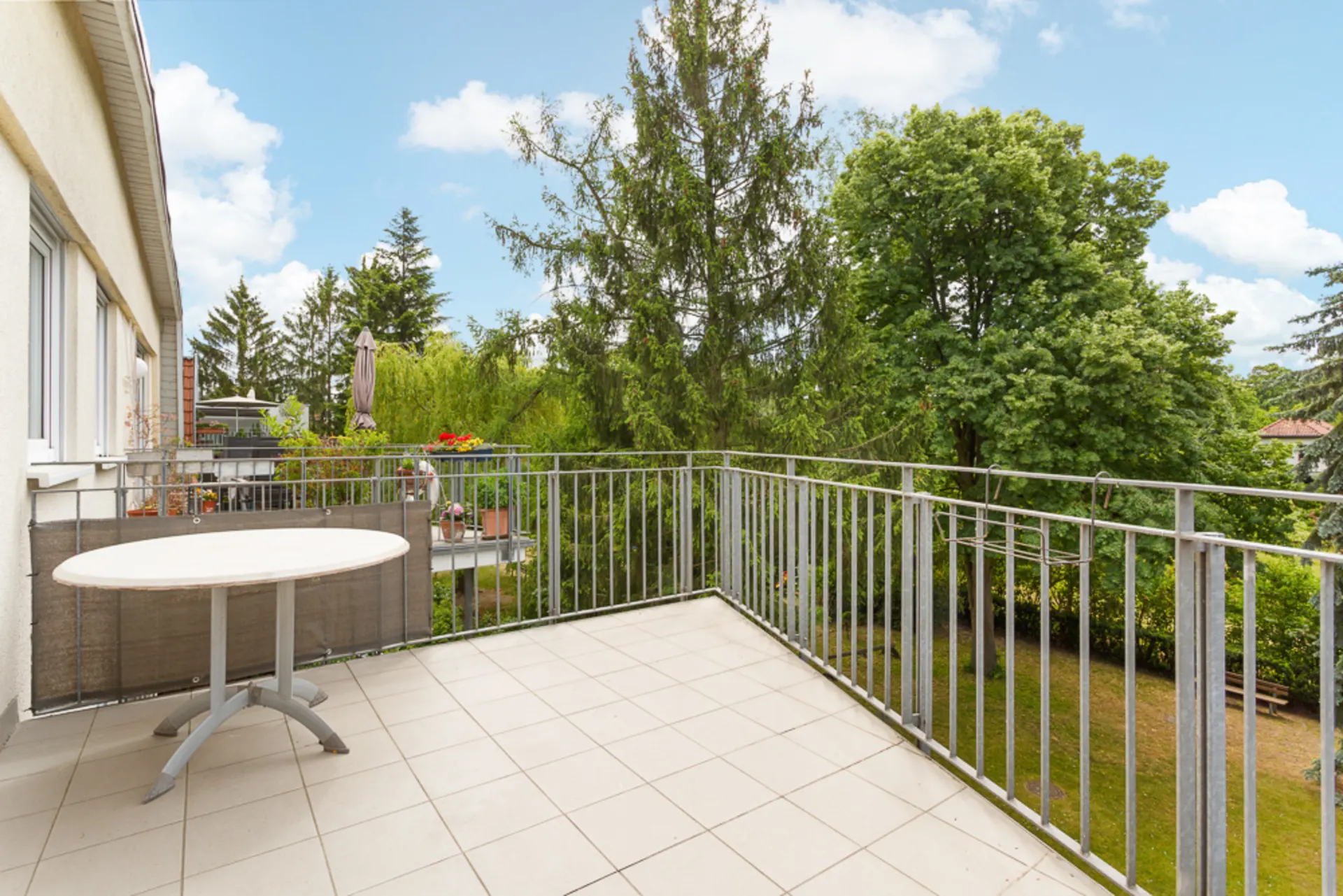 Helle Balkonterrasse mit Metallgeländer, einem runden Tisch und Blick auf grüne Bäume.