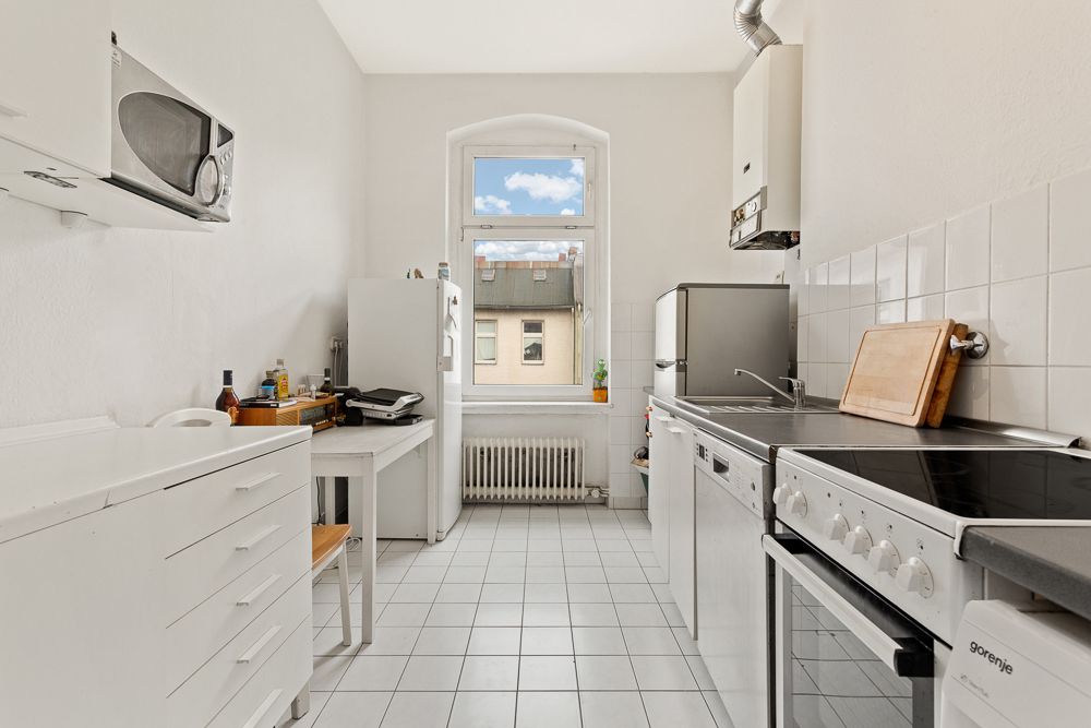 Helle Küche mit weißen Fliesen, Herd, Kühlschrank, Mikrowelle und Fenster mit Blick nach draußen.