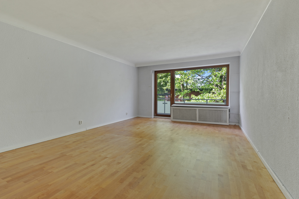 Leeres Wohnzimmer mit Holzboden und großem Fenster, das auf einen Balkon mit Blick ins Grüne führt.