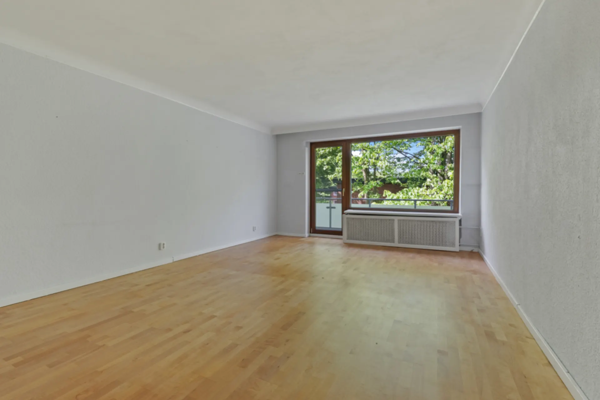Leeres Wohnzimmer mit Holzboden und großem Fenster, das auf einen Balkon mit Blick ins Grüne führt.