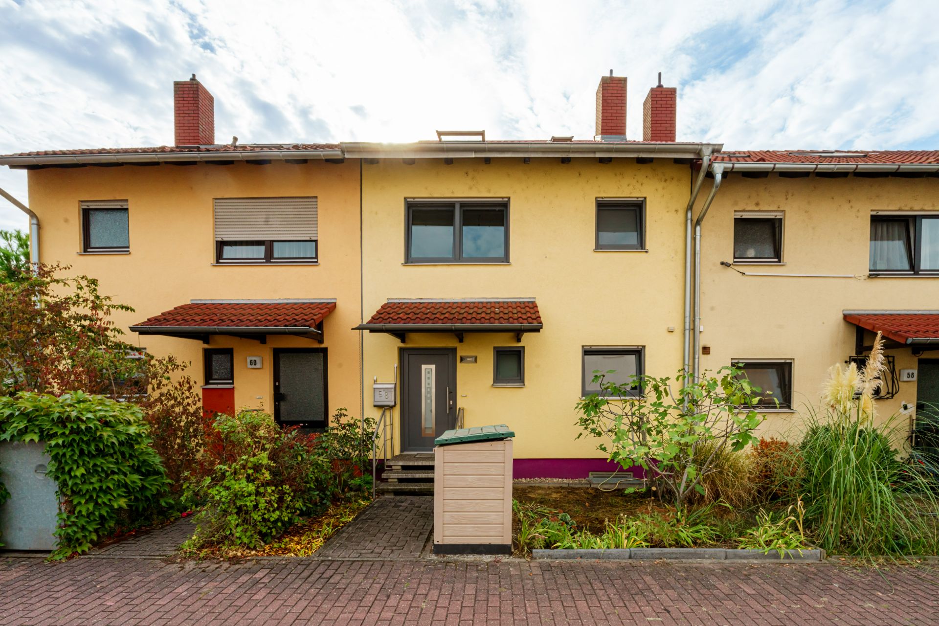 Reihenhaus mit gelber Fassade, Vorgarten und rotem Ziegeldach, neben gepflastertem Weg.