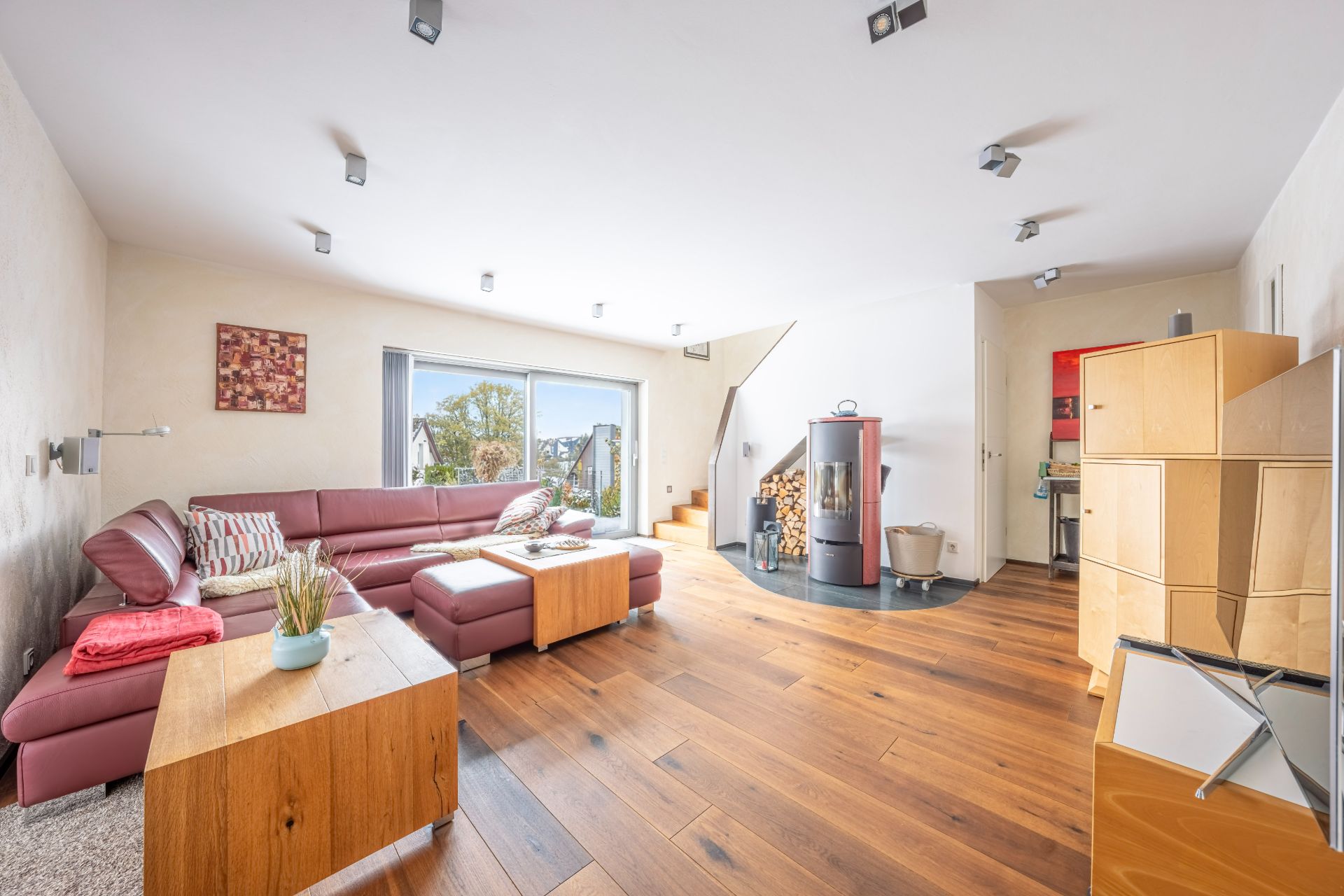 Helles Wohnzimmer mit Ecksofa, Kaminofen, Holzboden und großen Fenstern zum Garten.