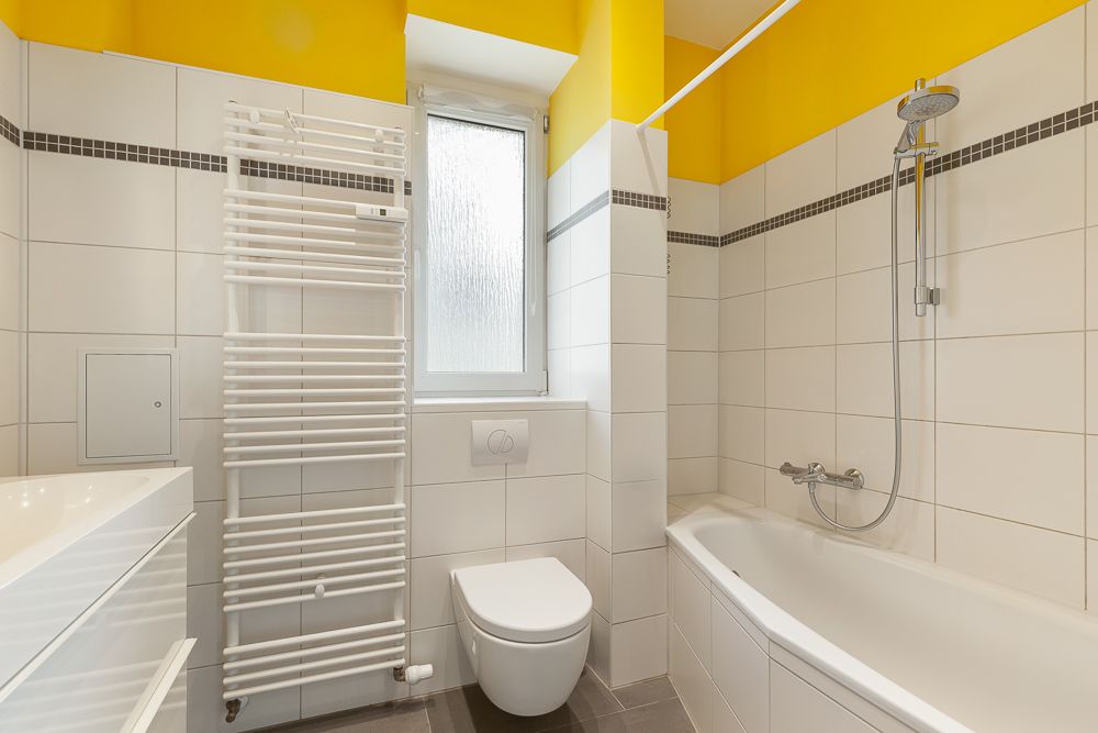 Modernes Badezimmer mit Badewanne, WC, Handtuchheizkörper und Fenster, weiße Fliesen und gelbe Wand.