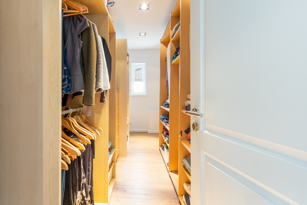 Ein begehbarer Kleiderschrank mit Holzregalen, Kleidung und Bügeln, beleuchtet durch Tageslicht.