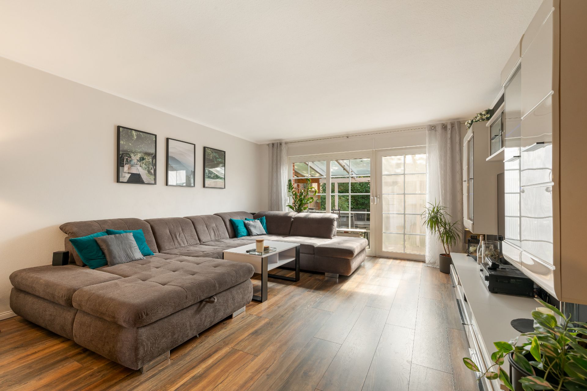 Helles Wohnzimmer mit großer Eckcouch, Couchtisch, Holzboden und Fensterfront mit Blick ins Grüne.