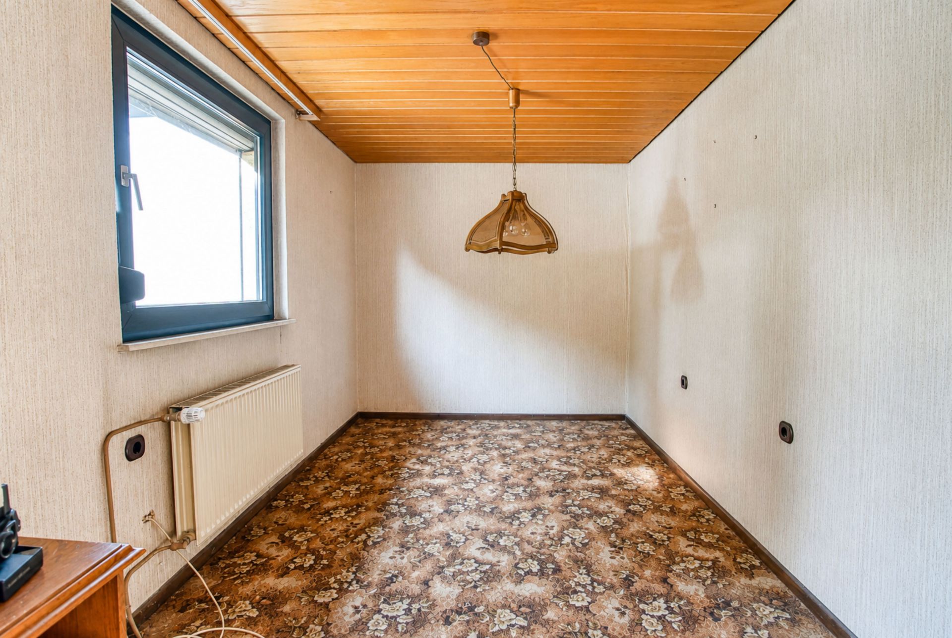Kleines, leeres Zimmer mit Teppichboden, Holzdecke, Fenster und Hängelampe.