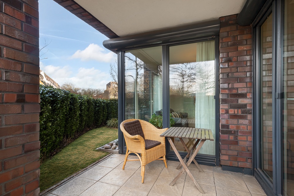 Kleine Terrasse mit Tisch, Stuhl und Blick auf Gartenhecke vor Glasfront.