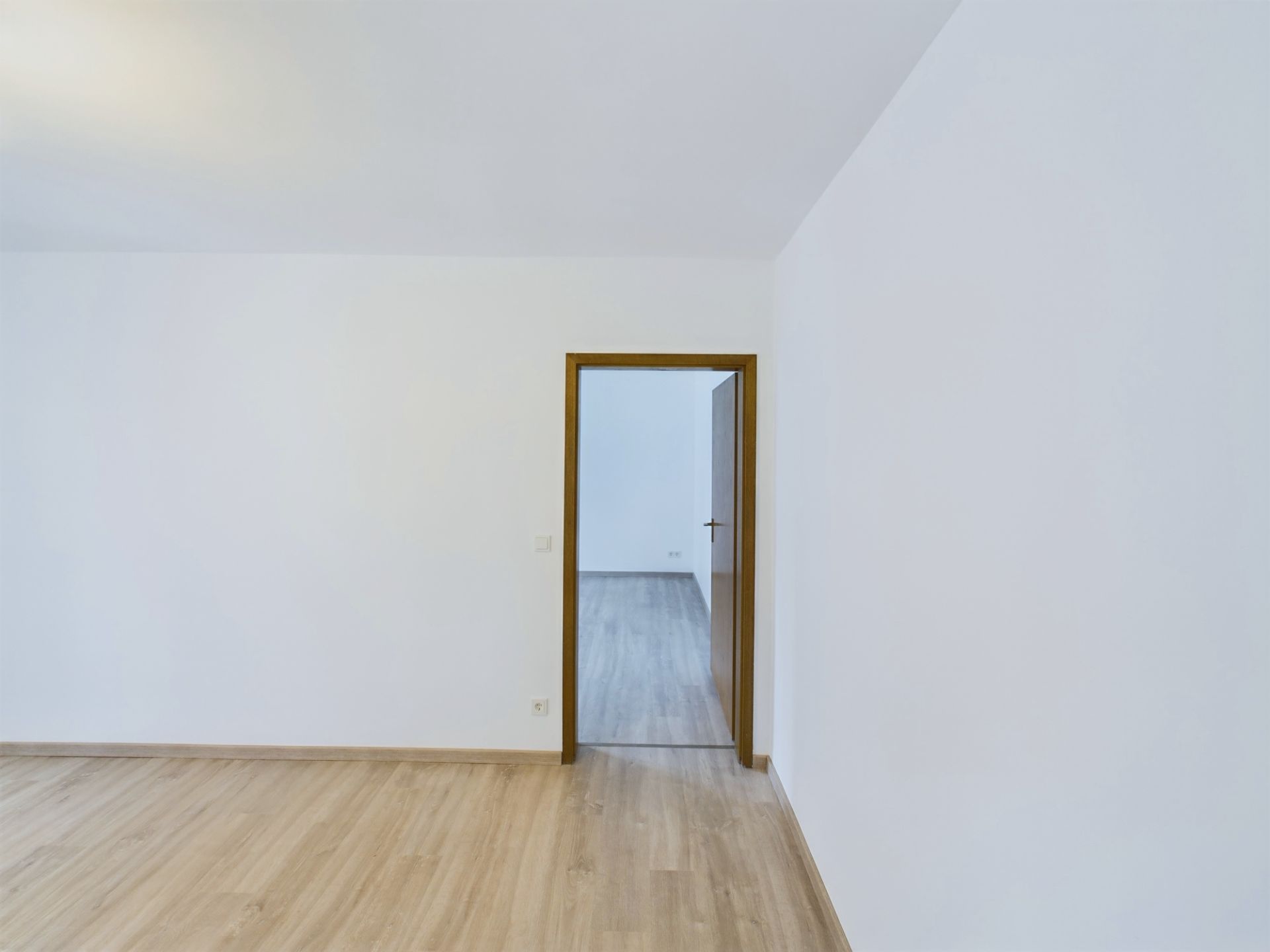 Leerer Raum mit Holzfußboden und weißer Wand, offene Tür führt zu weiterem Zimmer.