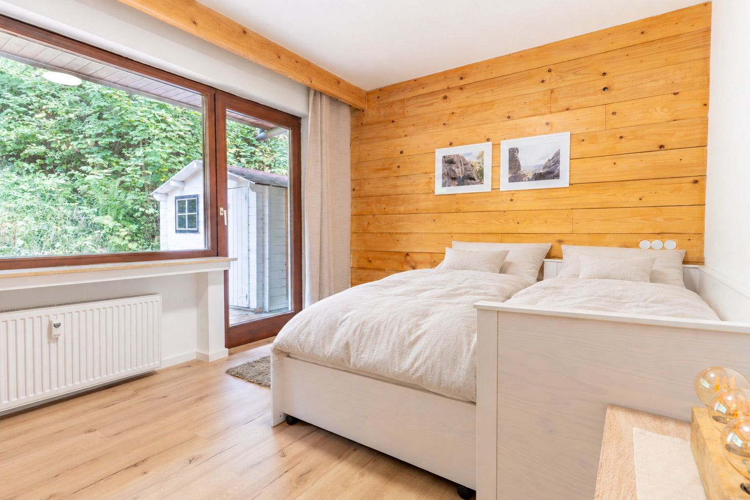 Helles Schlafzimmer mit zwei Betten, Holzwand und großem Fenster mit Blick auf den Garten.