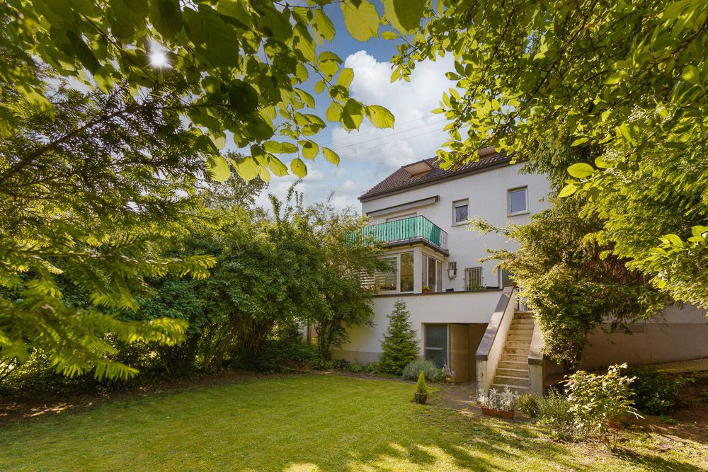 Haus mit Balkon, Außentreppe und grünem Garten, umgeben von Bäumen und Sträuchern.