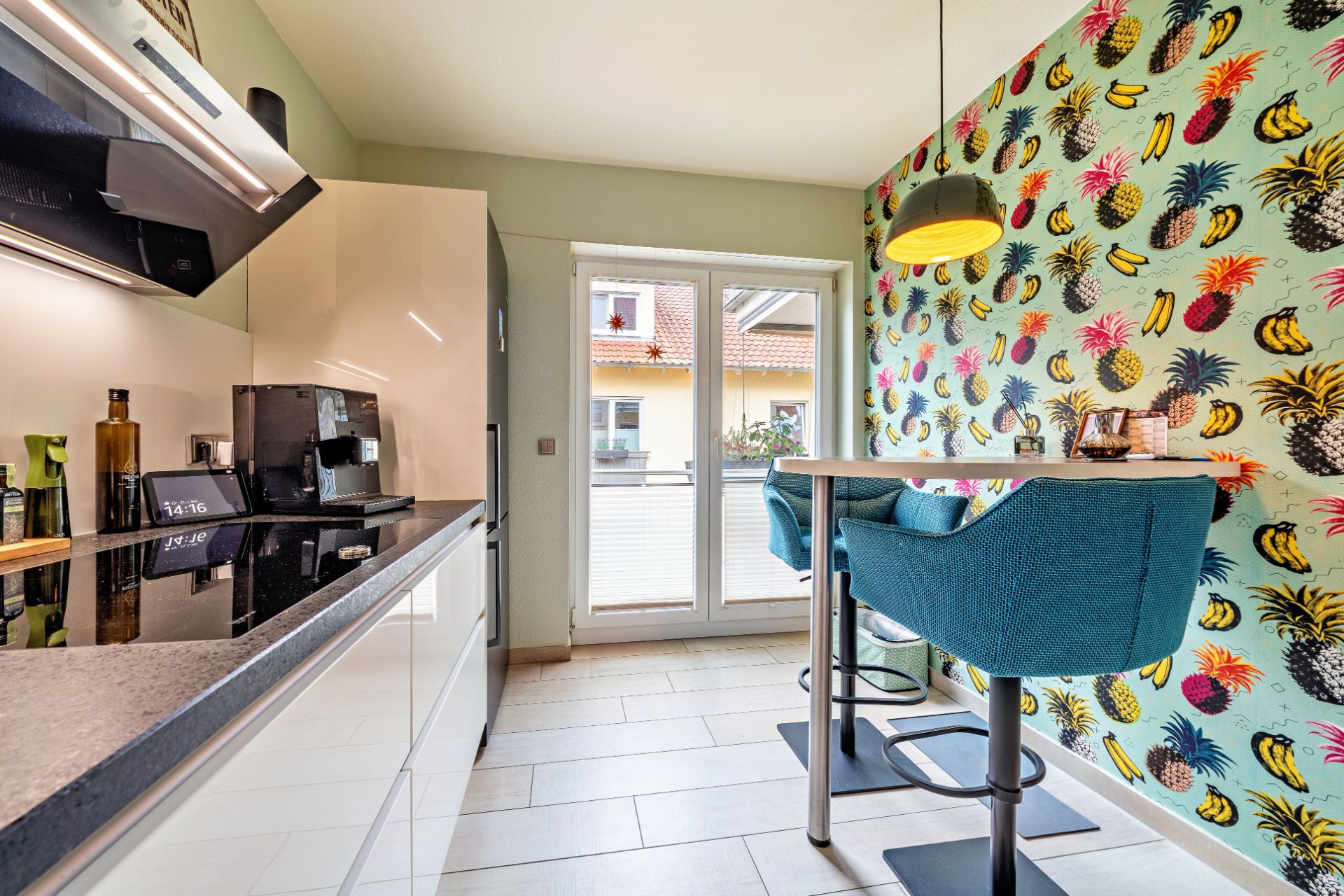 Helle Küche mit bunter Tapete, Bartisch und zwei blauen Stühlen vor Glastür zum Balkon.