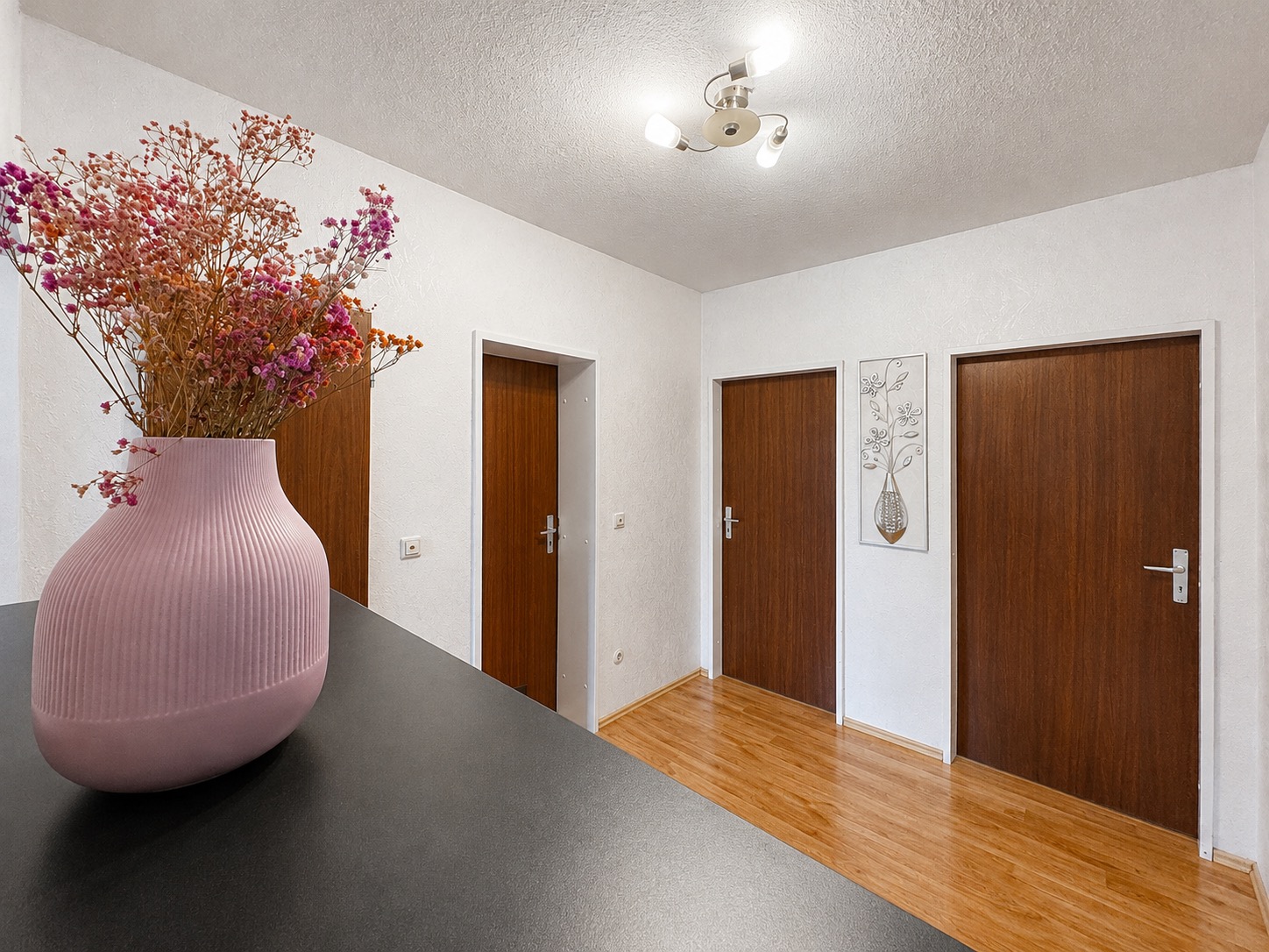 Flur mit drei braunen Türen, Holzfußboden, rosa Vase mit Trockenblumen im Vordergrund.