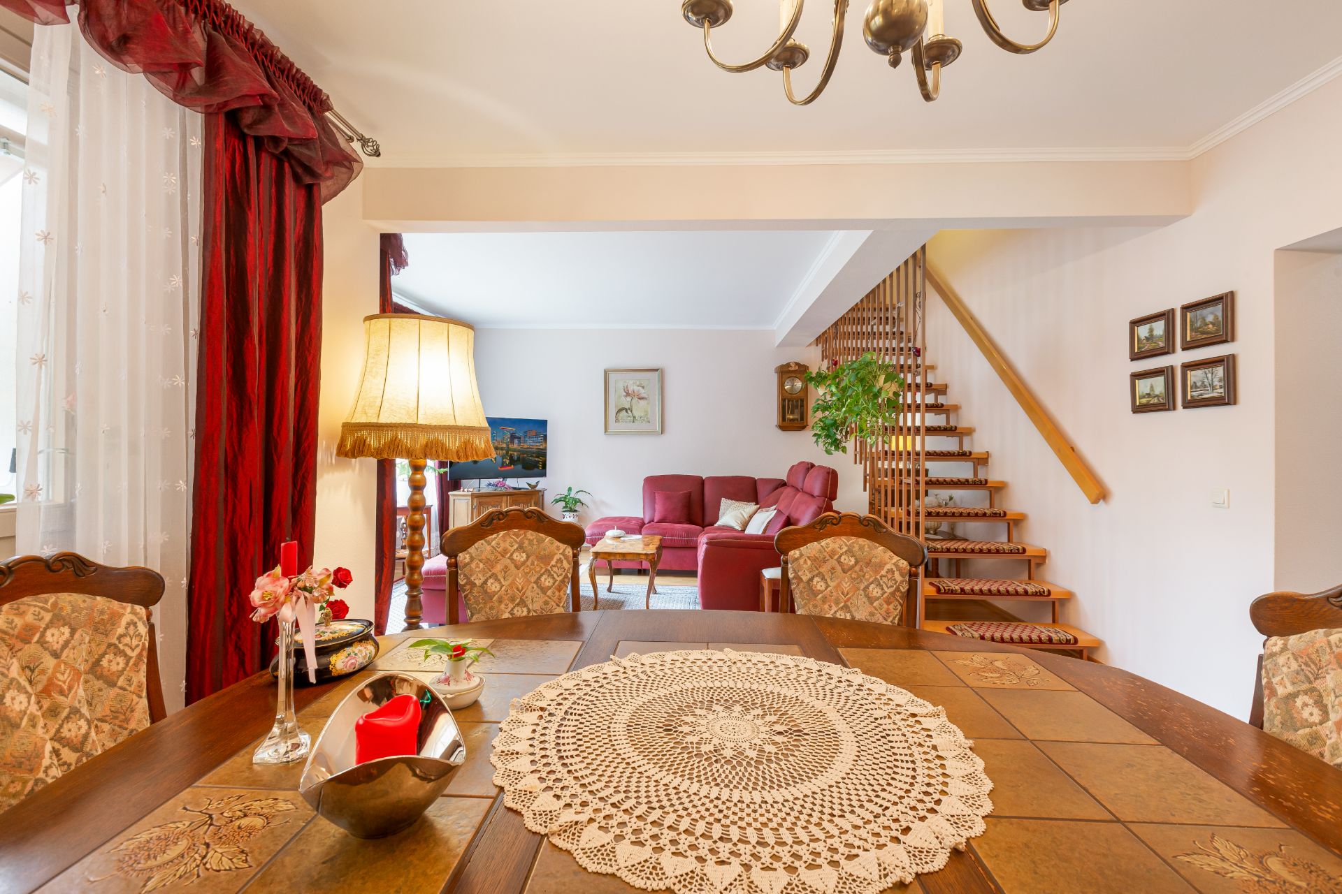 Esszimmer mit Holztisch und gehäkelter Decke, dahinter Wohnzimmer mit rotem Sofa und Holztreppe.