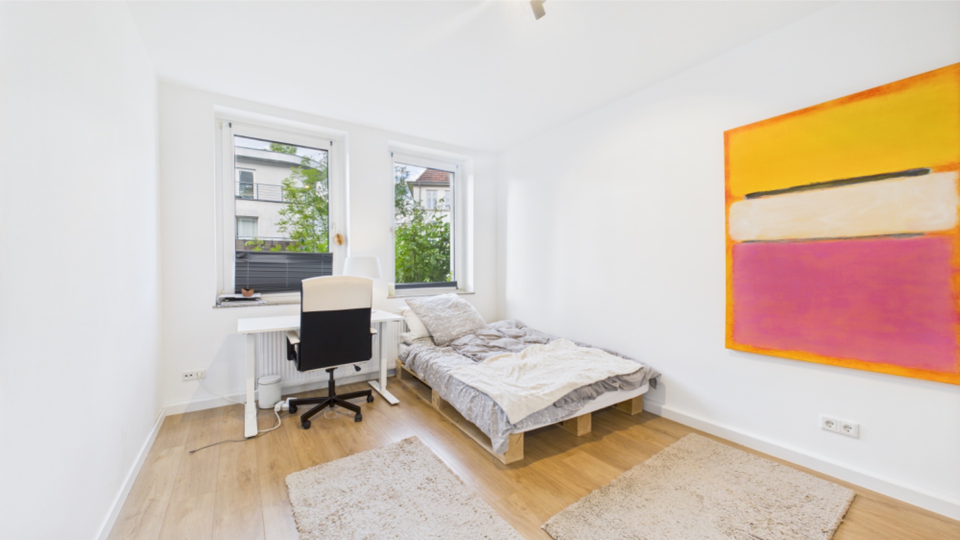 Helles Schlafzimmer mit Einzelbett, Schreibtisch am Fenster und großem bunten Bild an der Wand.