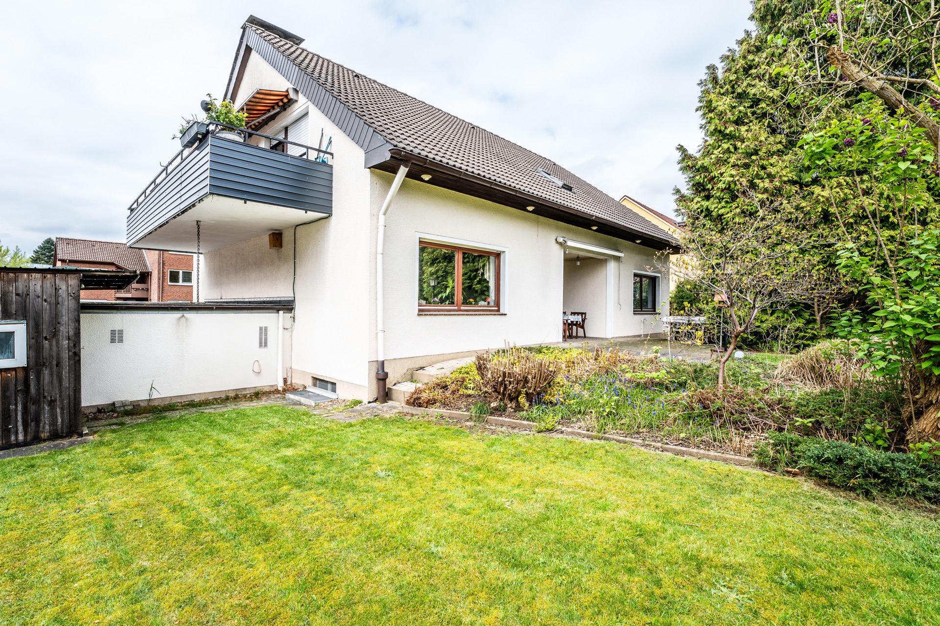 Einfamilienhaus mit Balkon, Terrasse und Garten, umgeben von Bäumen und Rasen.
