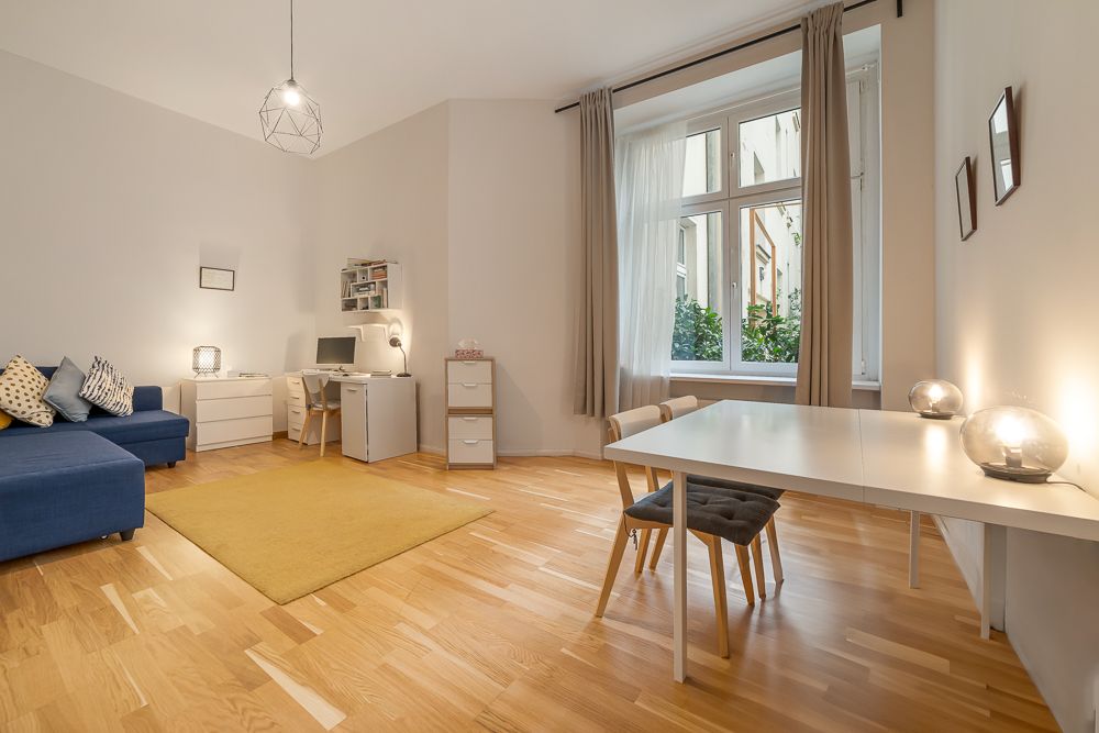 Helles Wohnzimmer mit Ecksofa, Schreibtisch, Esstisch und großem Fenster mit Vorhängen.