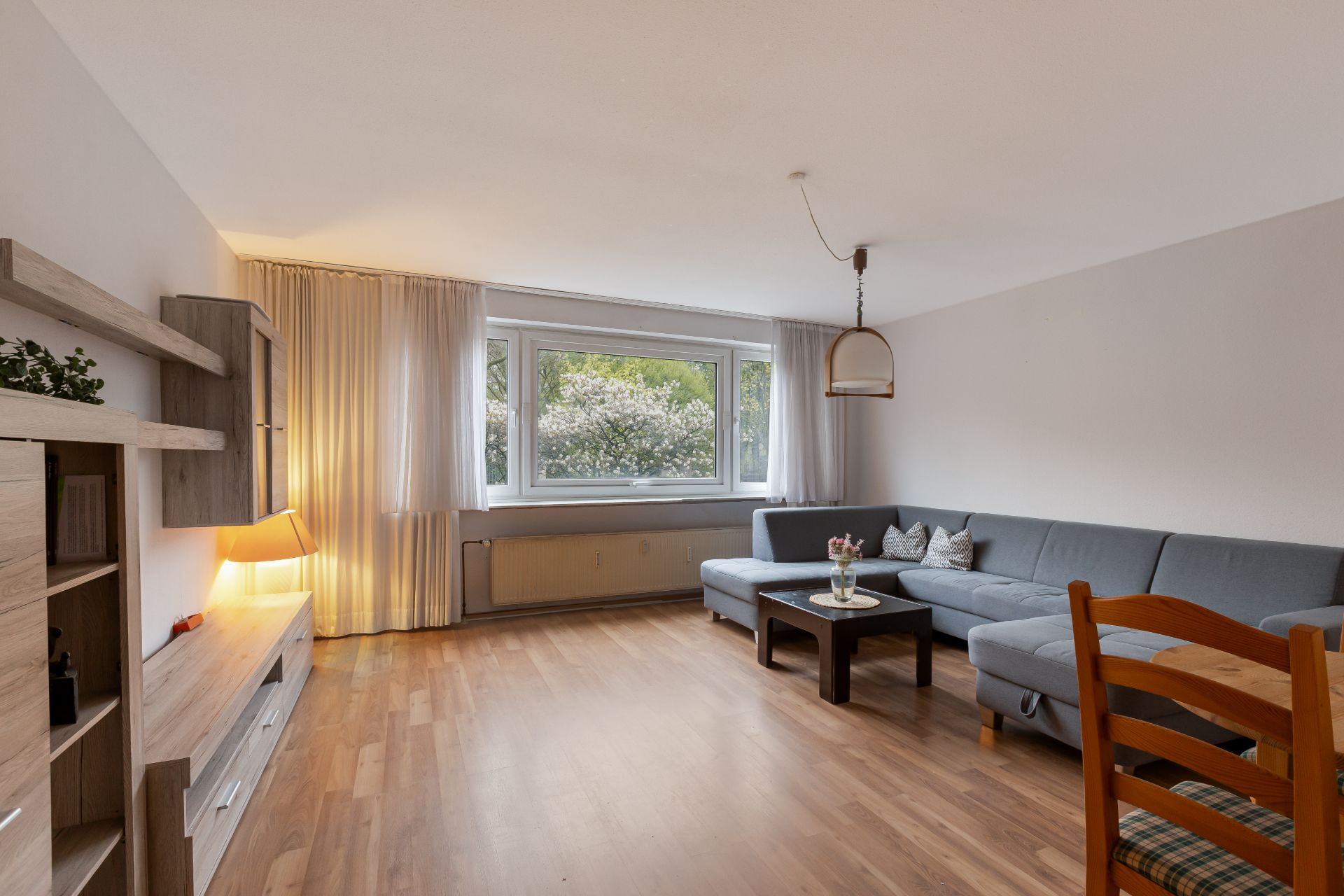 Helles Wohnzimmer mit großem Fenster, grauem Ecksofa und Holzfußboden.