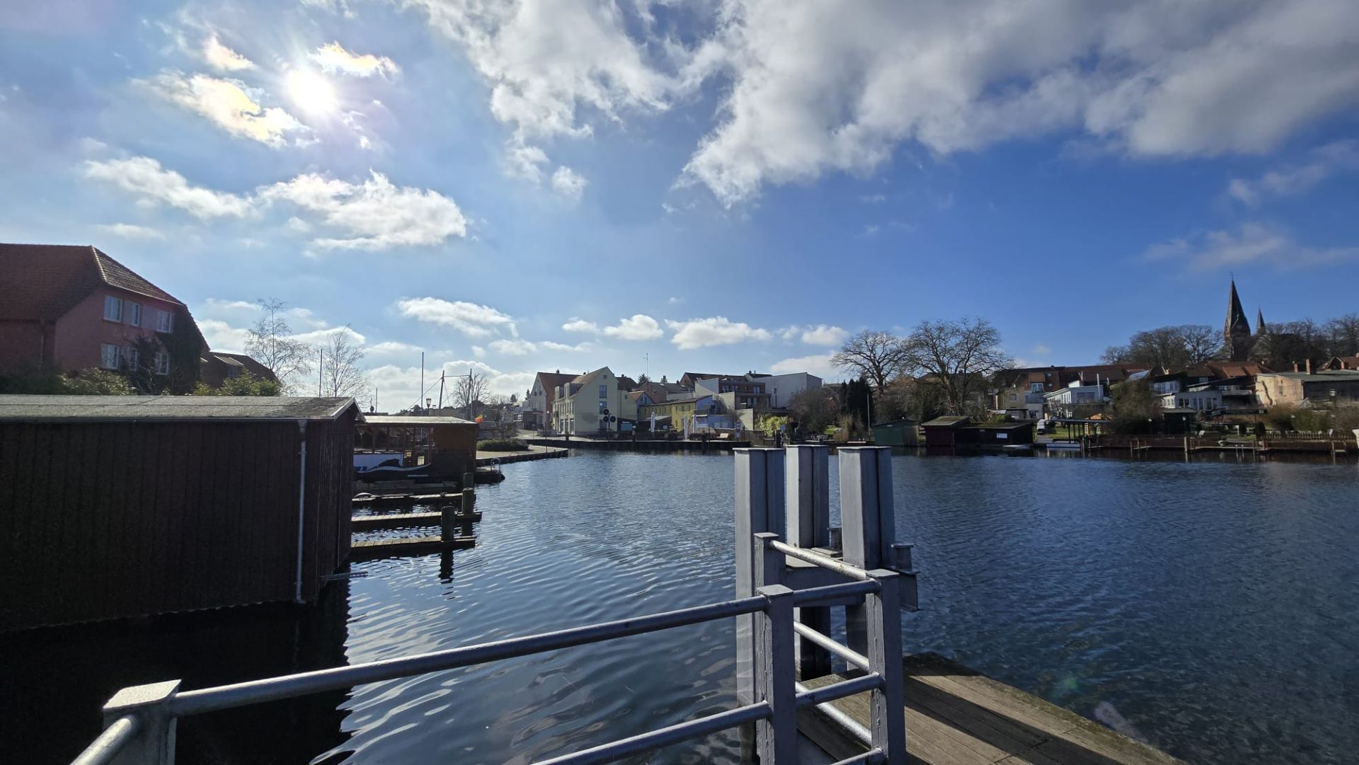 Blick auf einen Fluss mit Steg und Wohnhäusern am Ufer unter blauem Himmel mit Wolken.