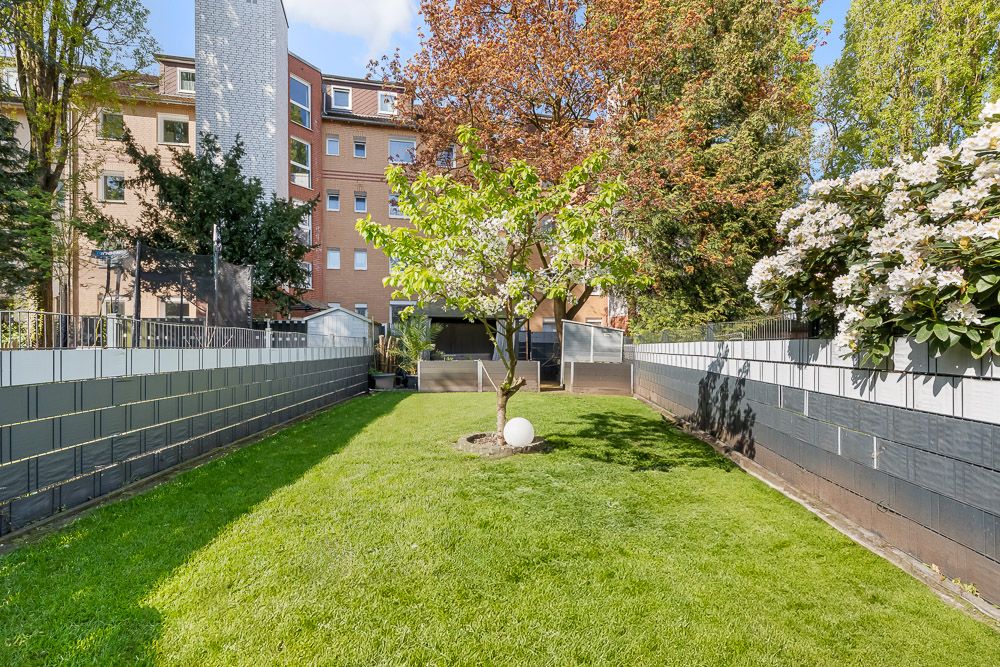 Gepflegter Garten mit grünem Rasen, mittigem Baum und angrenzenden Wohngebäuden im Hintergrund.