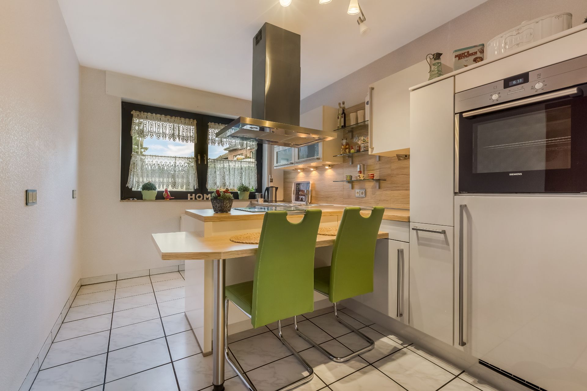 Moderne Küche mit Esstheke, zwei grünen Stühlen und Fenster mit Spitzenvorhängen.
