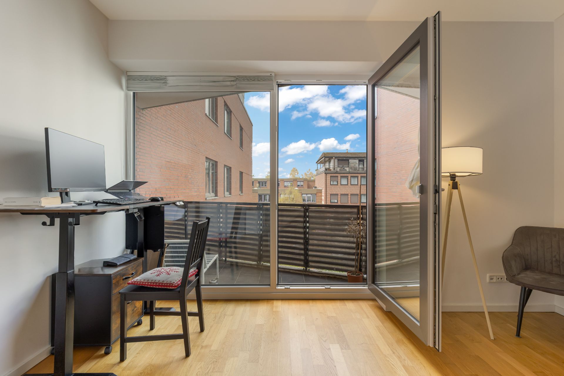 Helles Arbeitszimmer mit Schreibtisch, Stuhl, Stehlampe und Zugang zu einem Balkon mit Stadtblick.
