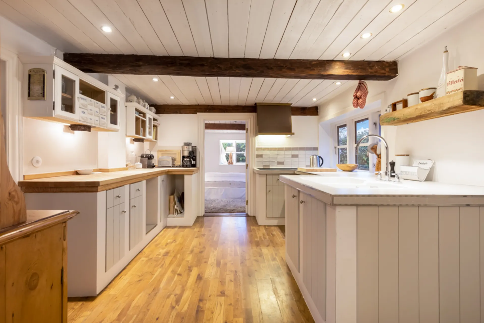 Helle Küche mit Holzboden, weißen Schränken, Holzbalken und Blick in ein Schlafzimmer.
