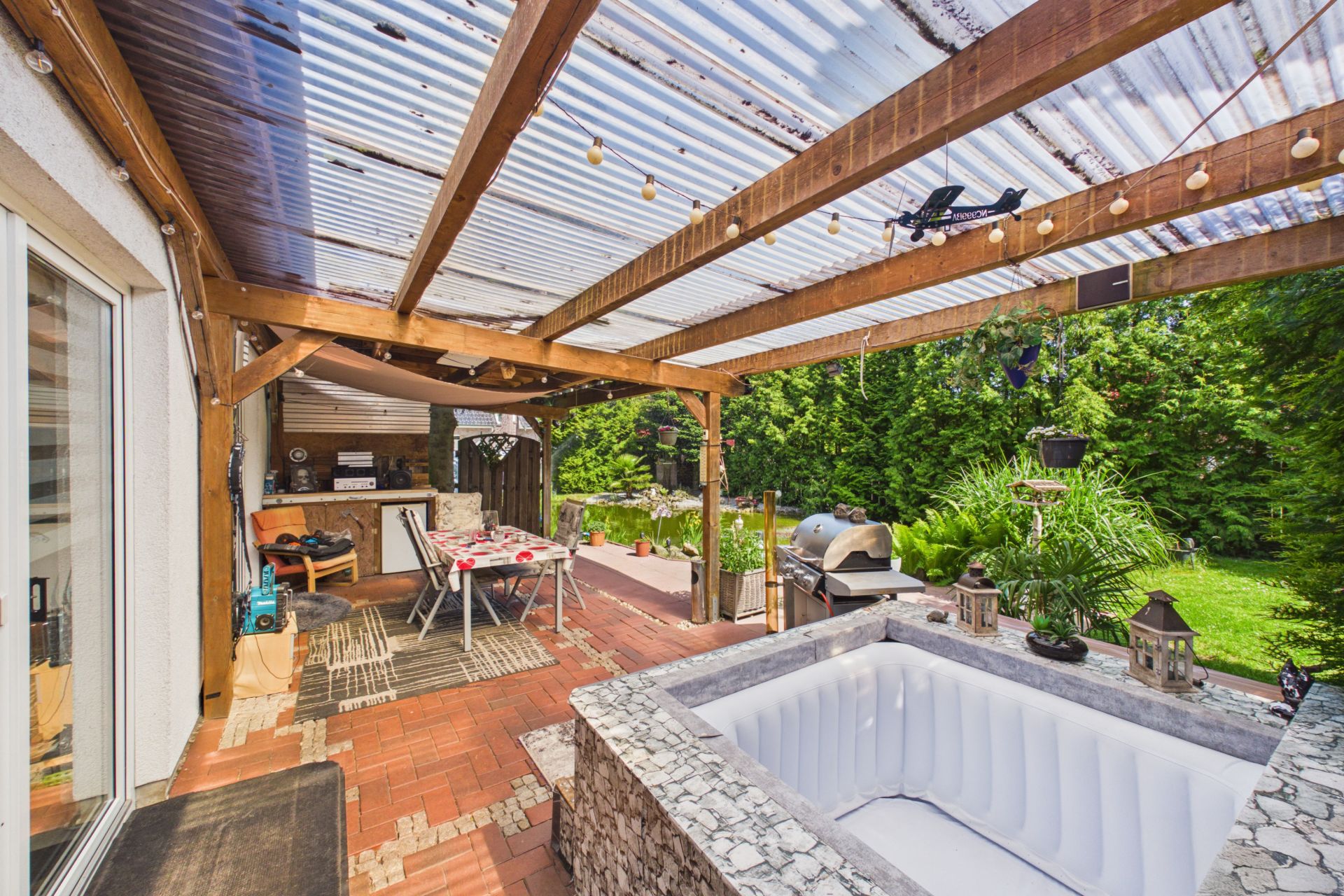 Überdachte Terrasse mit aufblasbarem Pool, Grill, Tisch und Blick in den Garten.