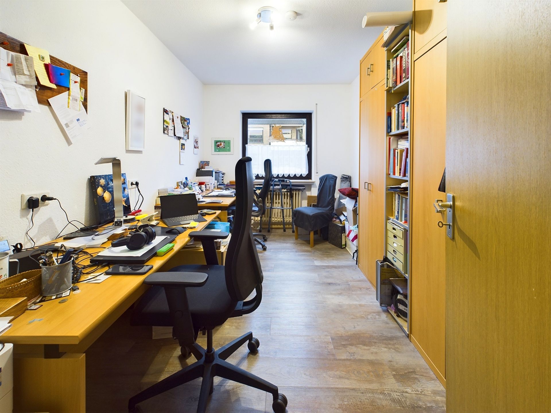 Helles Arbeitszimmer mit zwei Schreibtischen, Bürostühlen, Regalen und einem Fenster am Ende des Raumes.