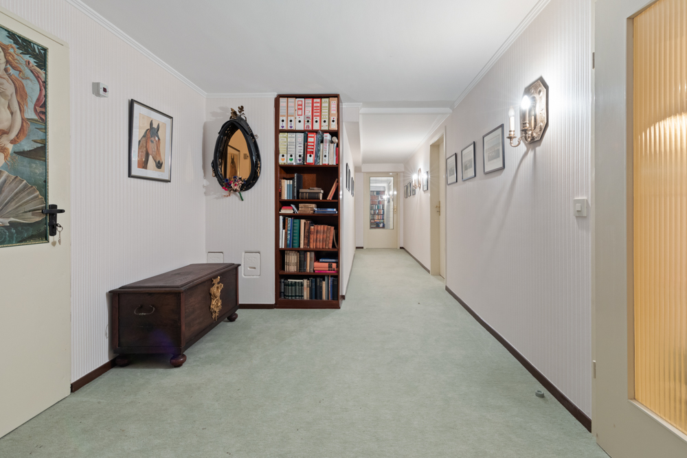 Langer Flur mit Teppich, Bücherregal, Holzkiste und gerahmten Bildern an den Wänden.