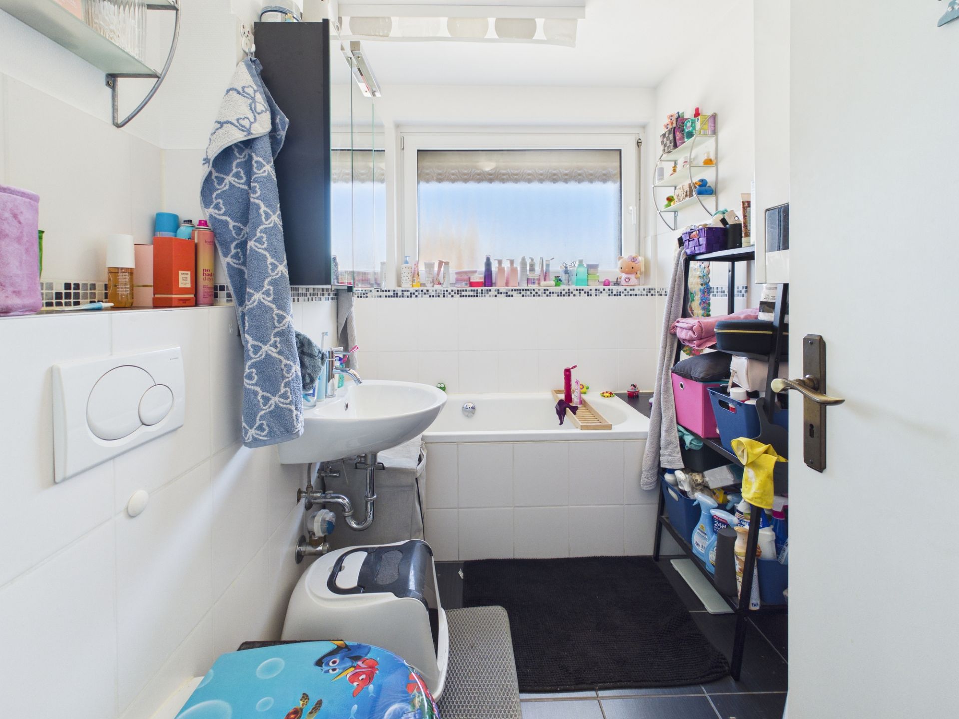 Kleines Badezimmer mit Badewanne, Waschbecken, Regal und verschiedenen Pflegeprodukten.