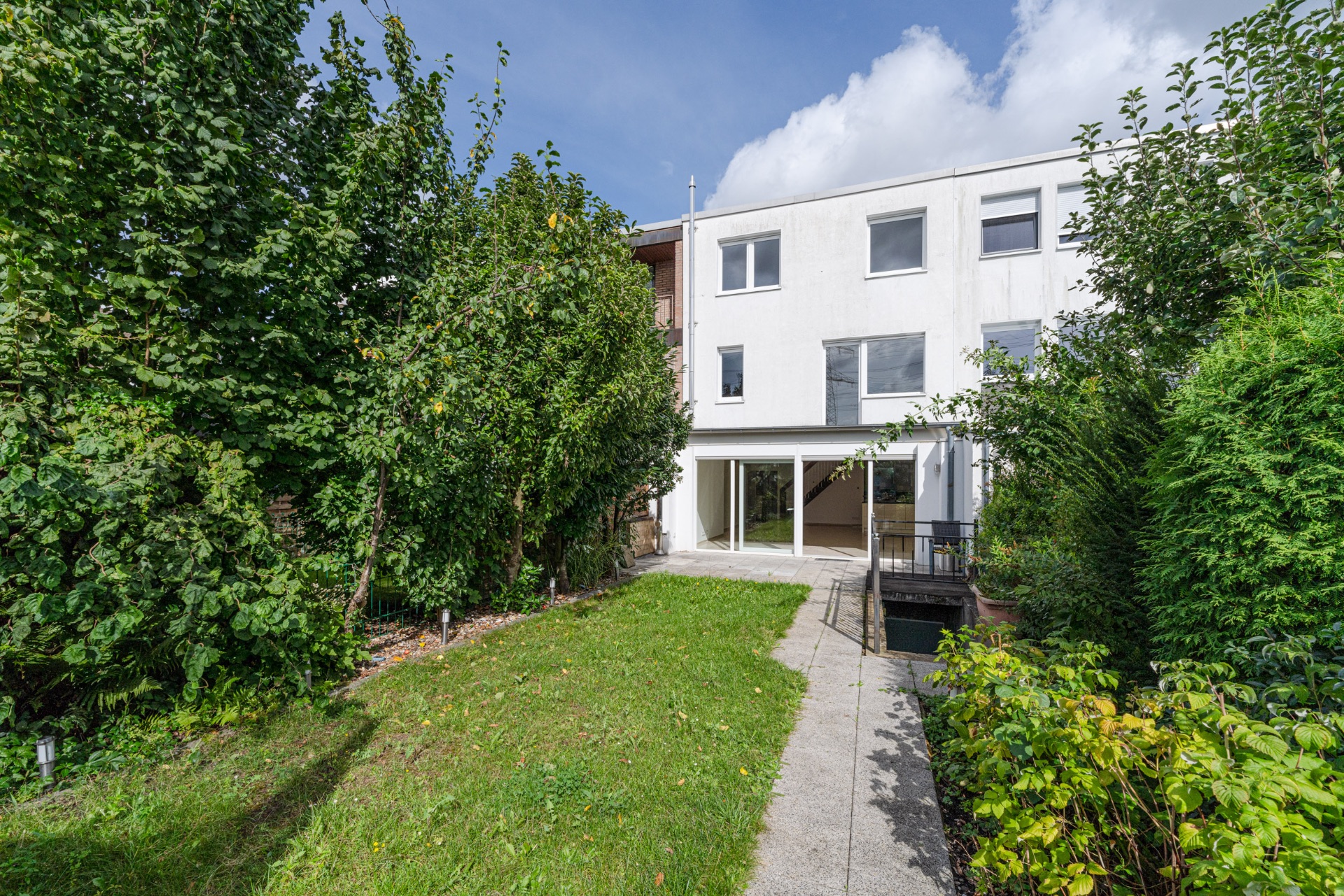 Garten mit Weg vor einem modernen, weißen Wohnhaus und vielen grünen Bäumen.