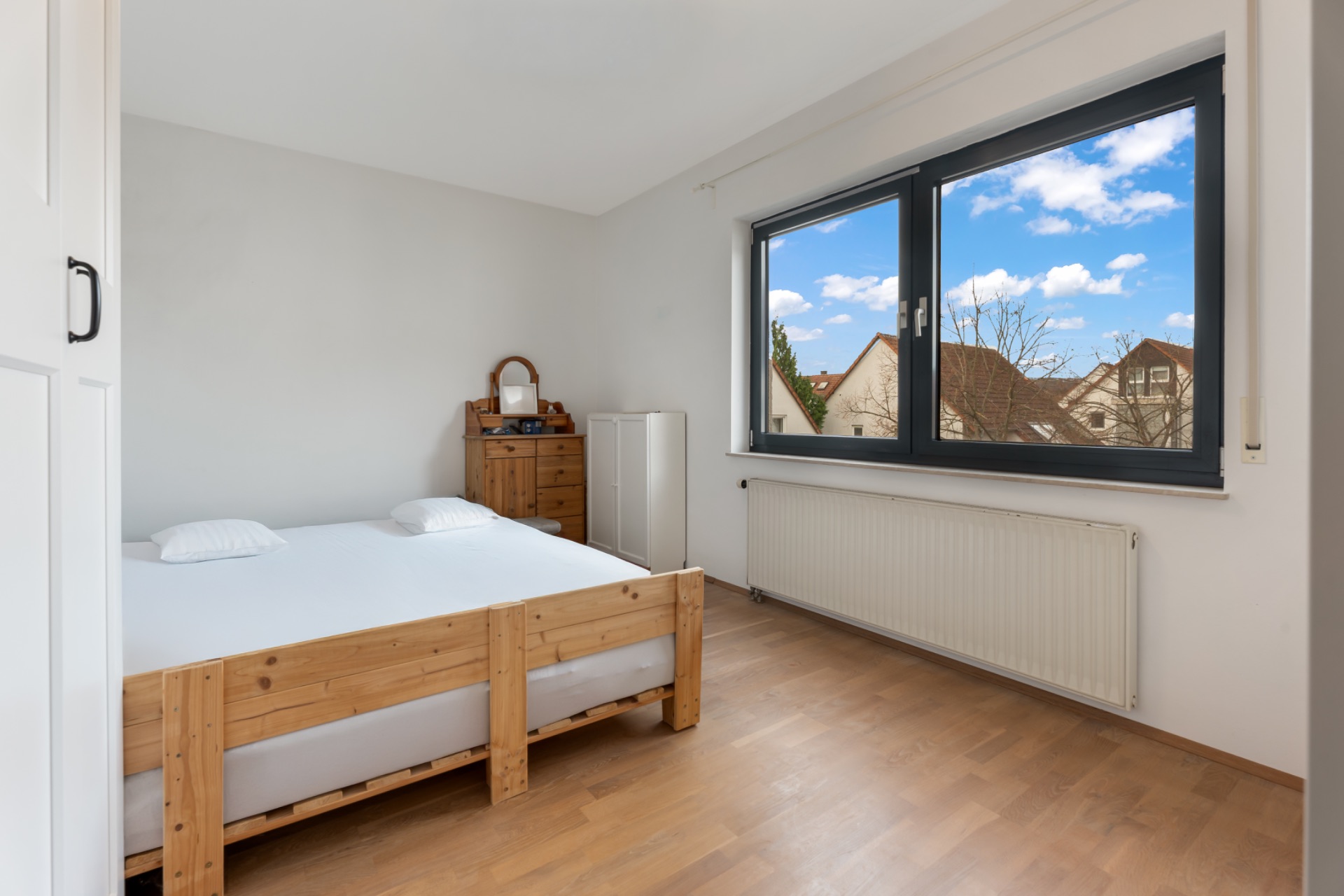 Helles Schlafzimmer mit Holzbett, großem Fenster und Kommode auf Parkettboden.