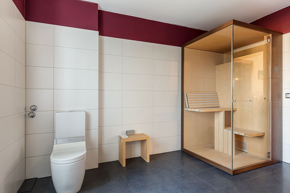Modernes Badezimmer mit Toilette, Holzbank und verglaster Sauna in einer Ecke.