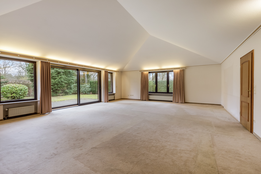 Großes, leeres Wohnzimmer mit Teppichboden, Fenstern und Glas-Schiebetüren zum Garten.