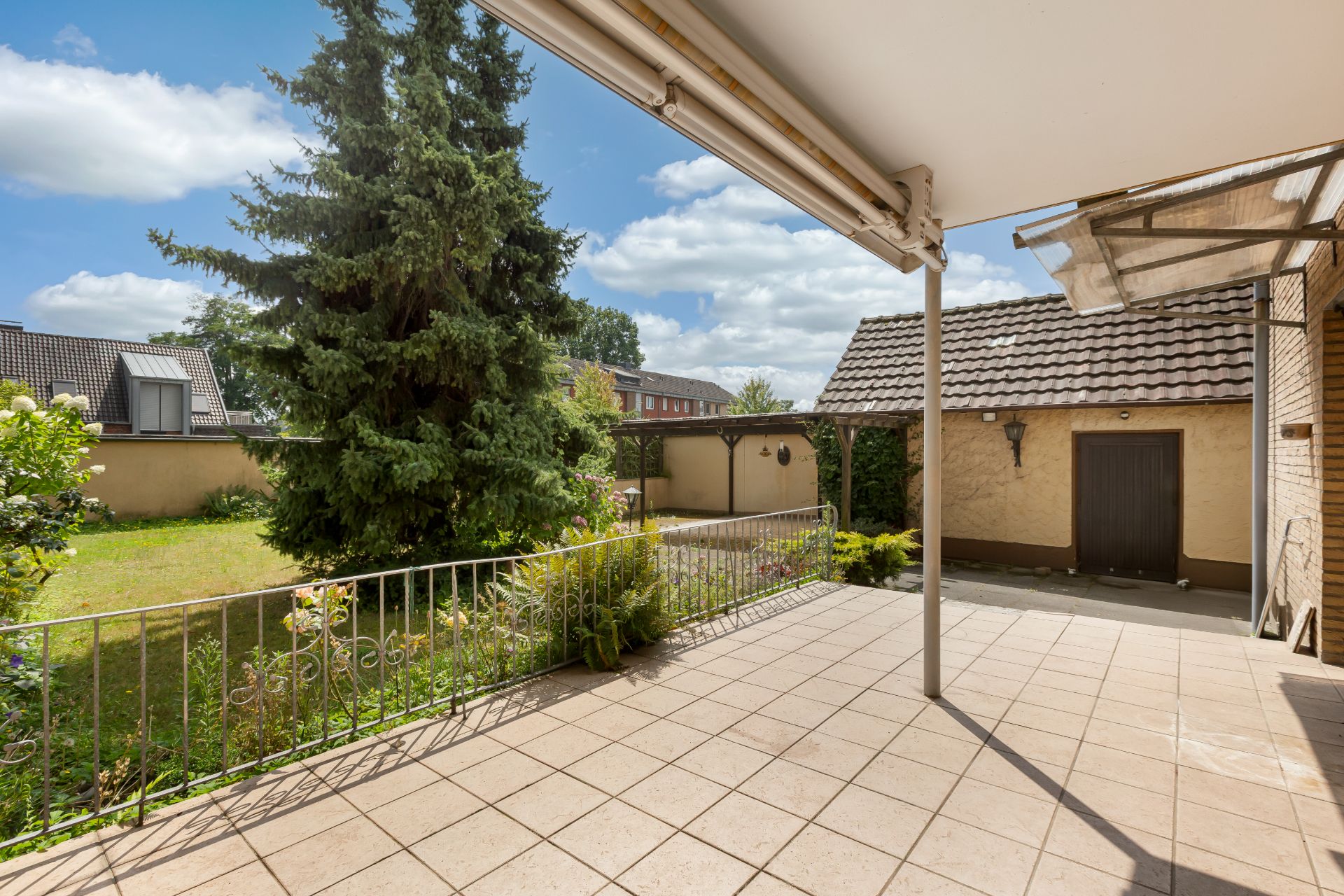 Überdachte Terrasse mit Fliesenboden, Blick auf Garten mit Baum und Nachbargebäude.