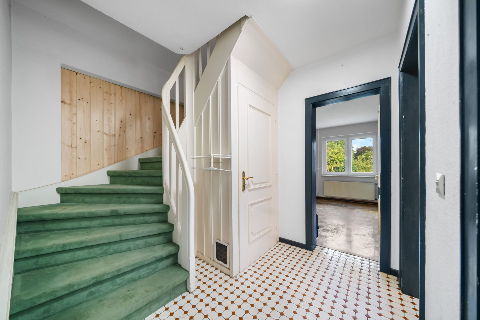 Flur mit gemustertem Boden, grüner Treppe und Zugang zu einem Zimmer mit großen Fenstern.