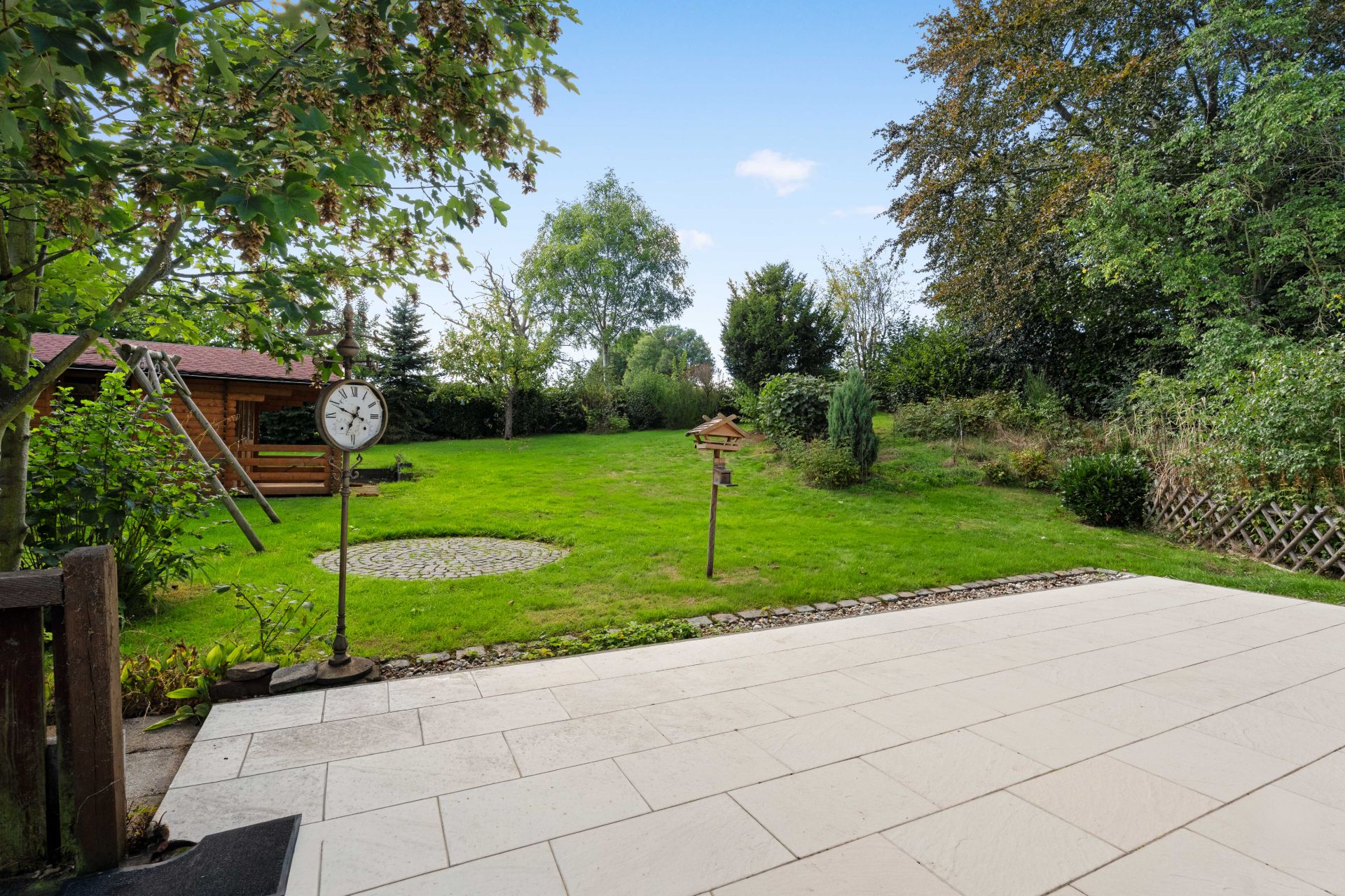 Blick von einer Terrasse auf einen großen grünen Garten mit Bäumen, Rasen und Vogelhaus.