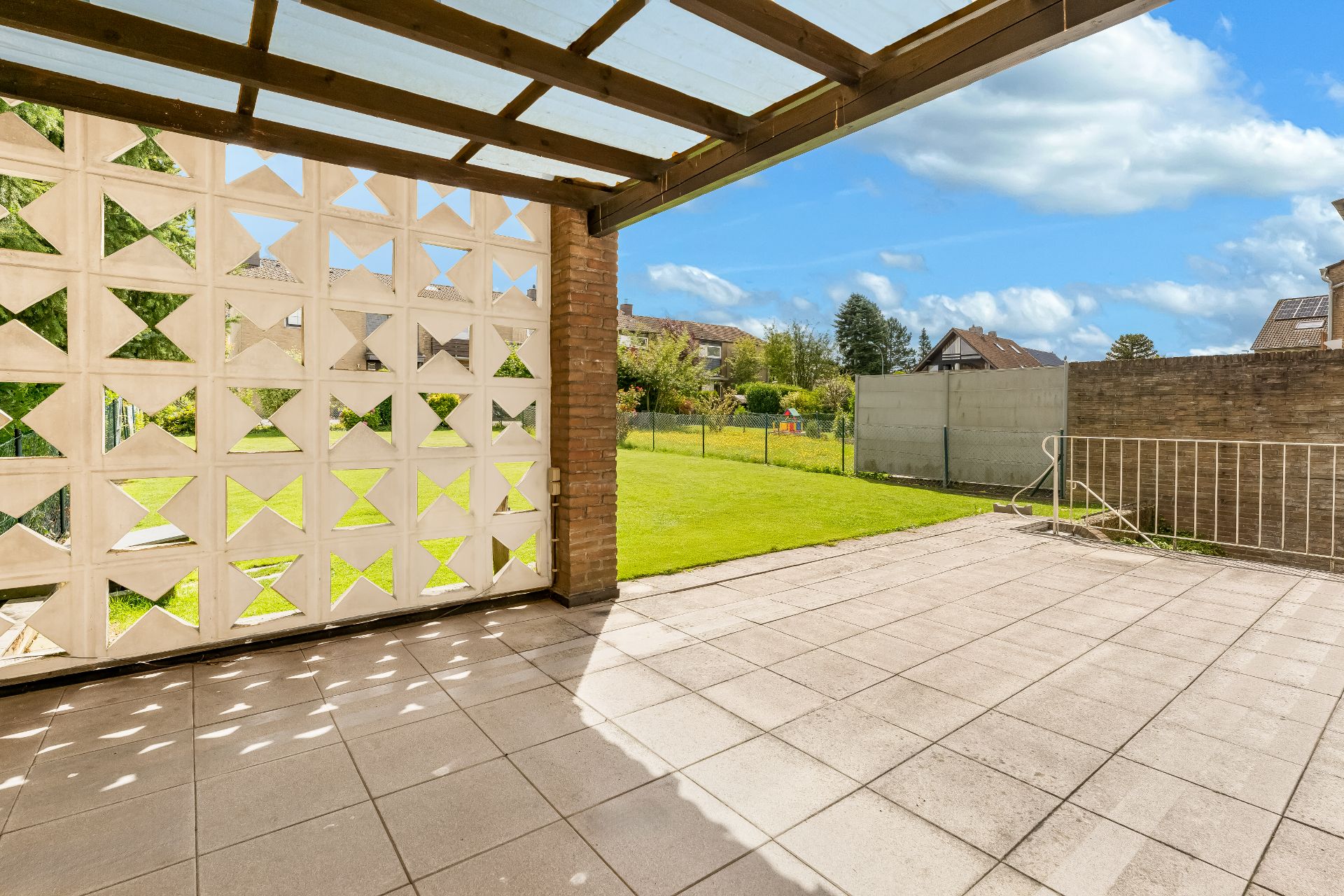 Überdachte Terrasse mit gemusterten Wandelementen und Blick auf einen Garten mit Rasen.