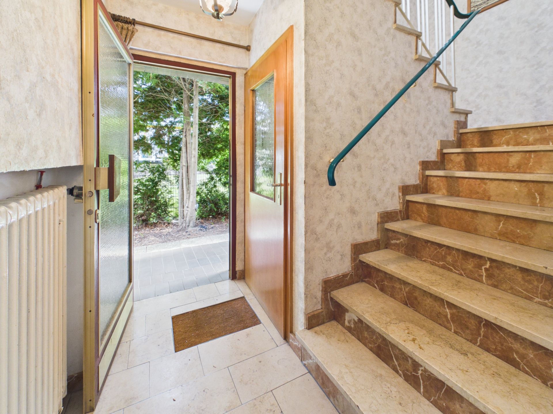 Hausflur mit offener Tür nach draußen und Treppe mit braunen Fliesen.
