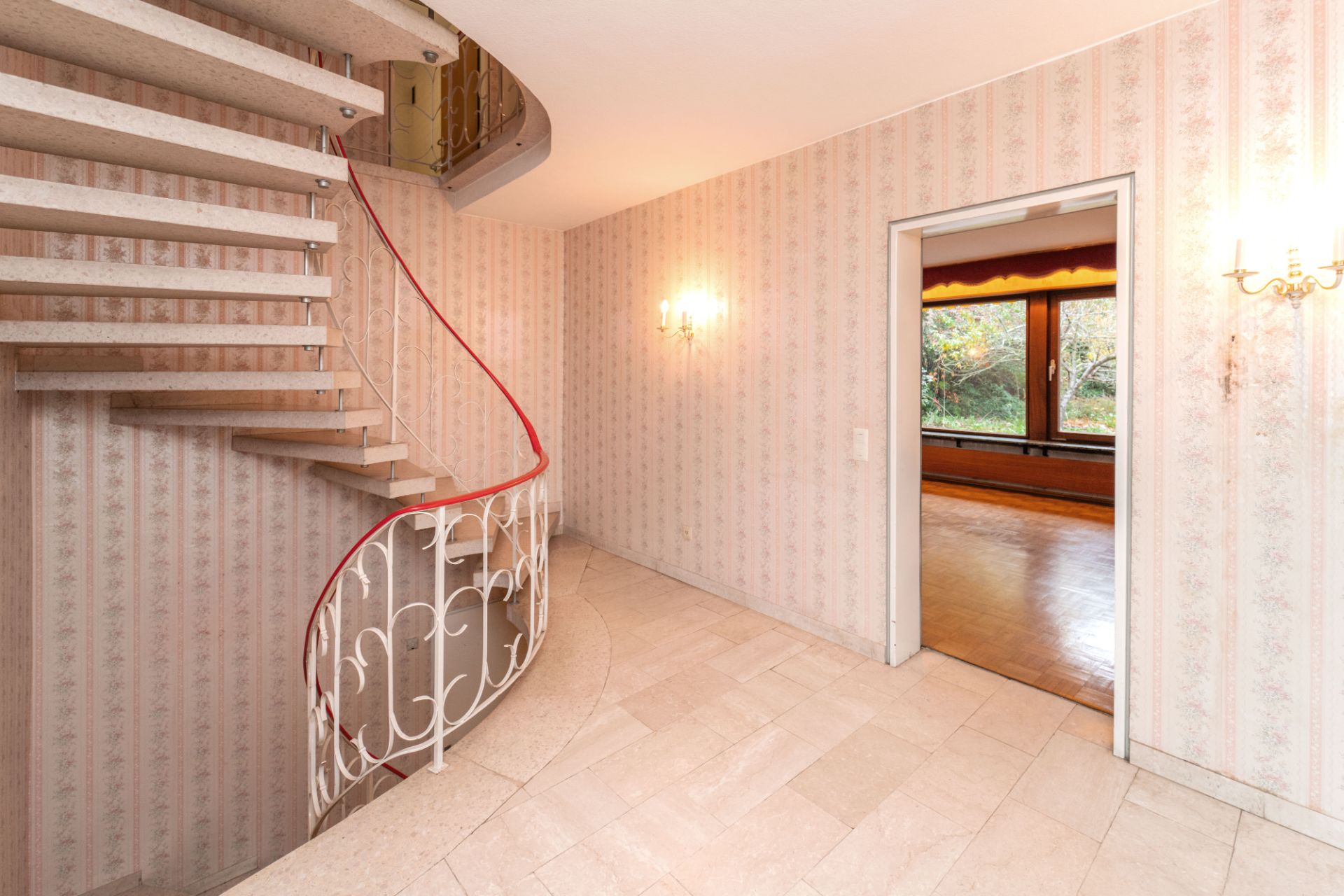 Flur mit Wendeltreppe, gemusterten Tapeten und Zugang zu einem Raum mit großen Fenstern.
