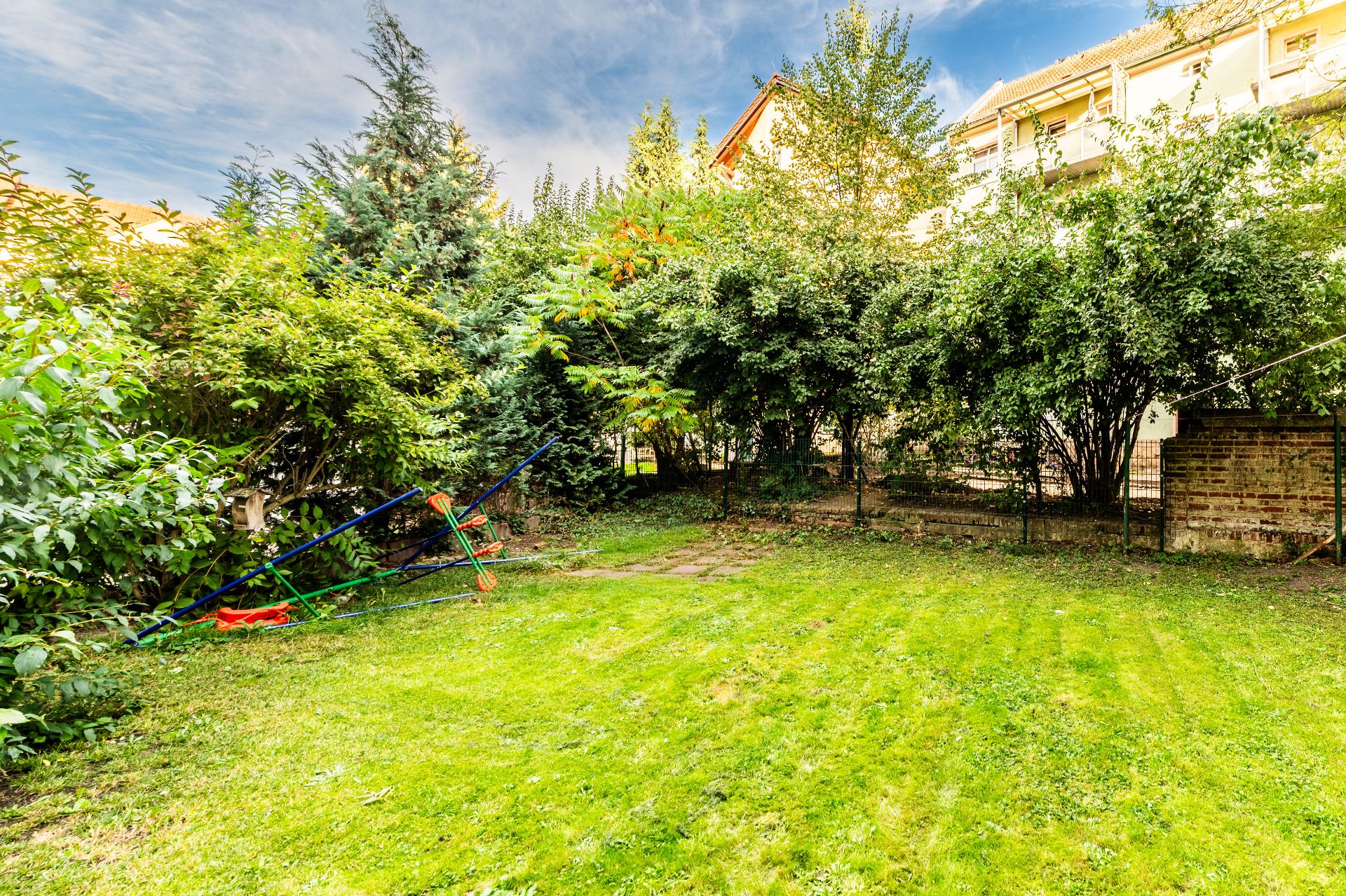 Grüner Garten mit Rasen, Bäumen und Sträuchern, im Hintergrund stehen Wohnhäuser.