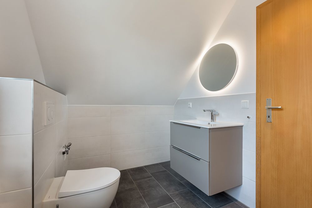 Modernes Badezimmer mit Wandtoilette, Waschbecken, rundem Spiegel und Dachschräge.
