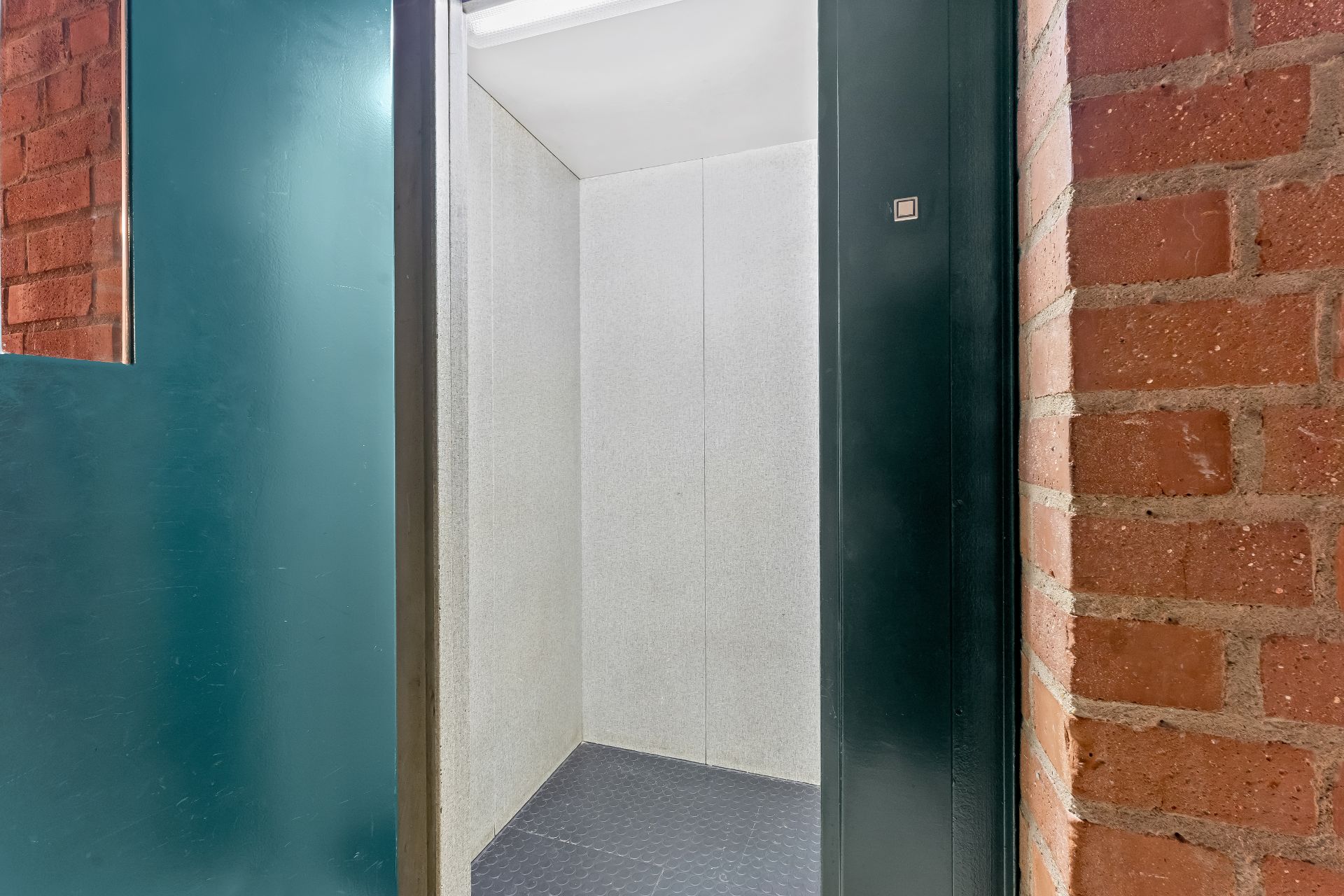 Offene Aufzugkabine mit grauen Wänden und dunklem Boden neben Backsteinwand.