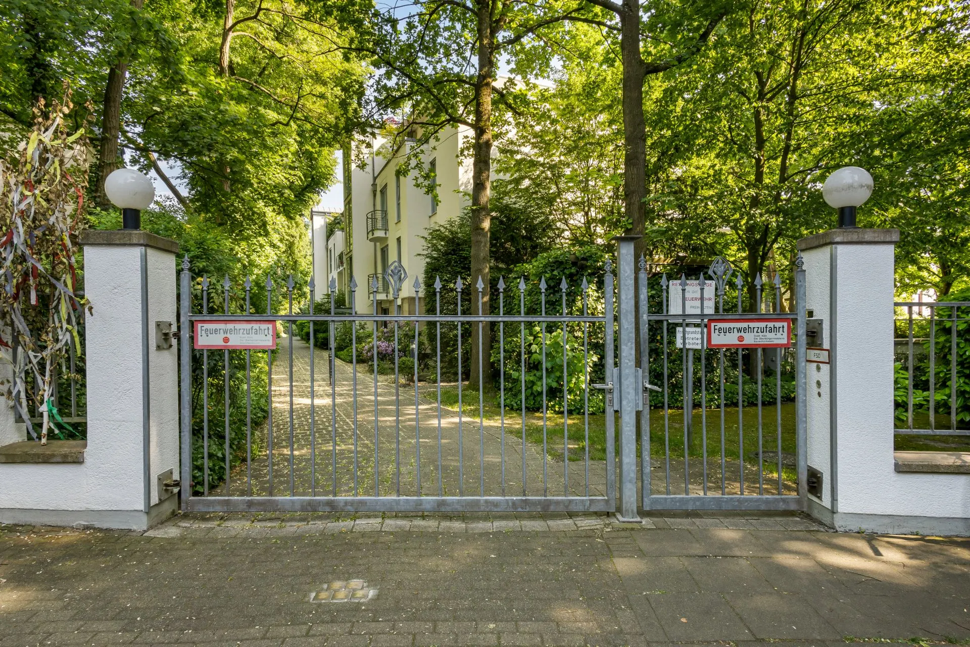 Metalltor mit zwei Säulen, dahinter Weg und Wohngebäude, umgeben von Bäumen und viel Grün.