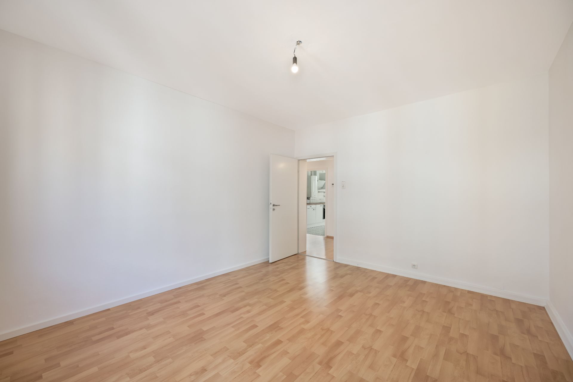 Heller, leerer Raum mit Holzboden, weißen Wänden und offener Tür zum nächsten Zimmer.