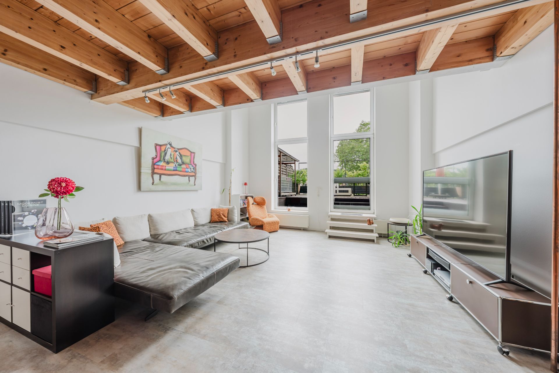 Modernes Wohnzimmer mit großen Fenstern, sichtbaren Holzbalken, Sofa, Sessel und Fernseher.