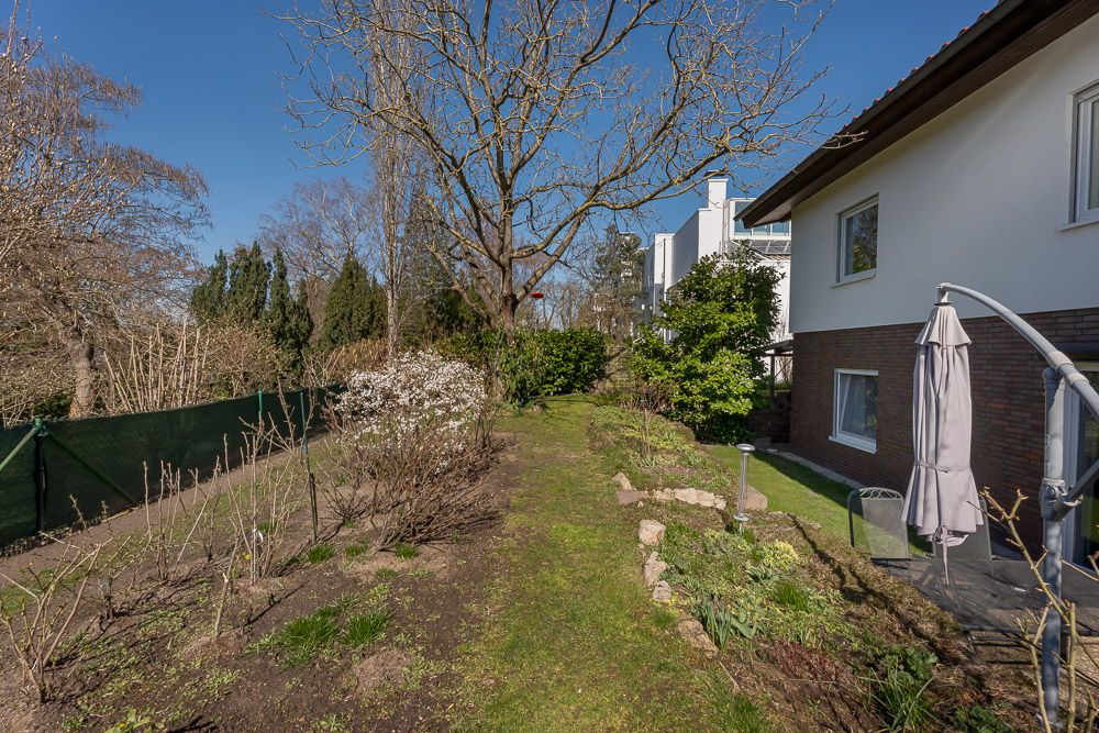 Garten mit Beeten, Bäumen und Terrasse neben einem Wohnhaus bei klarem Himmel.
