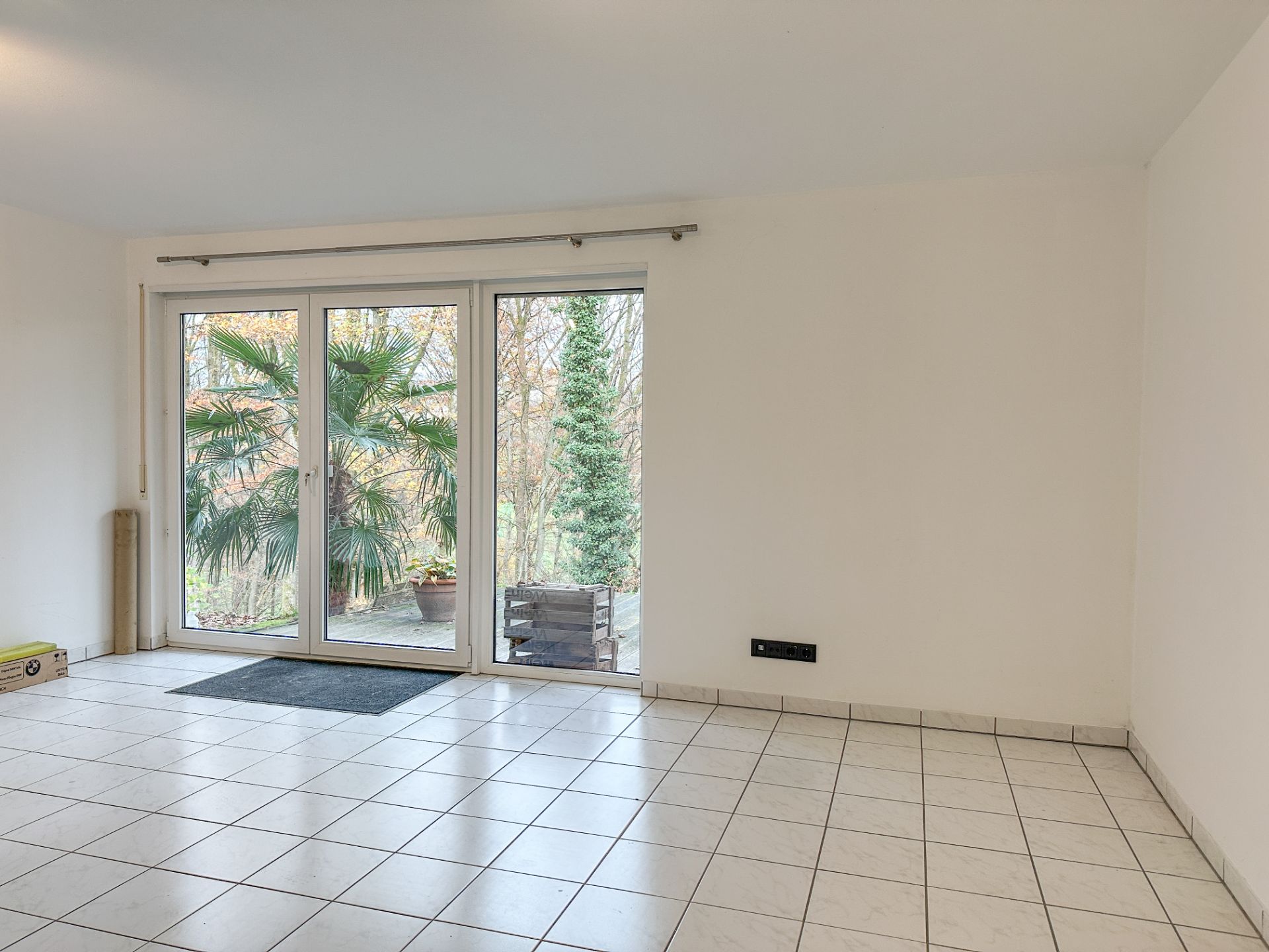 Helles Zimmer mit großen Glas-Schiebetüren und weiß gefliesten Boden, Blick auf Terrasse mit Pflanzen.